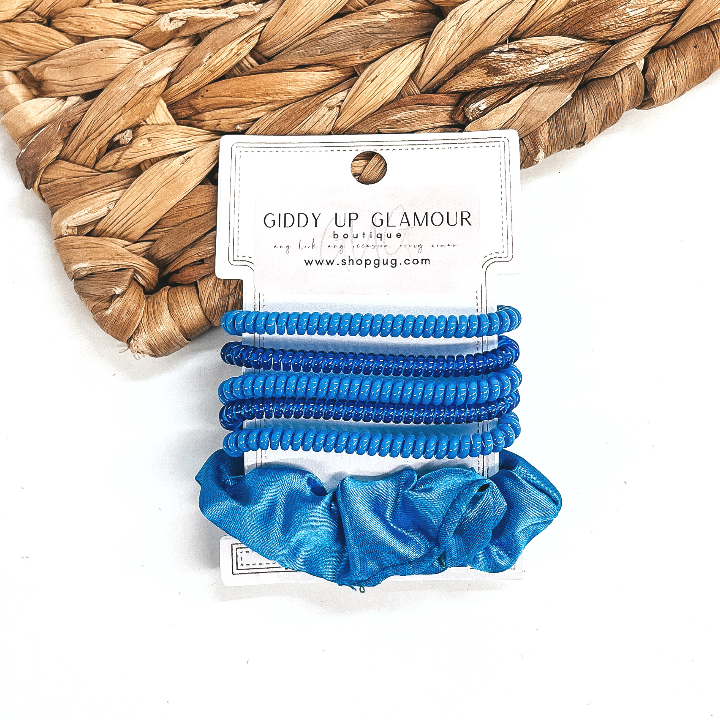 A set of six hair ties, five spiral and one satin scrunchie in blue. There are three  solid color spiral hair ties and two transparent. These hair ties are on a white  cardboard piece with a Giddy Up Glamour logo, they are leaned up against a brown woven  plate and a white background.