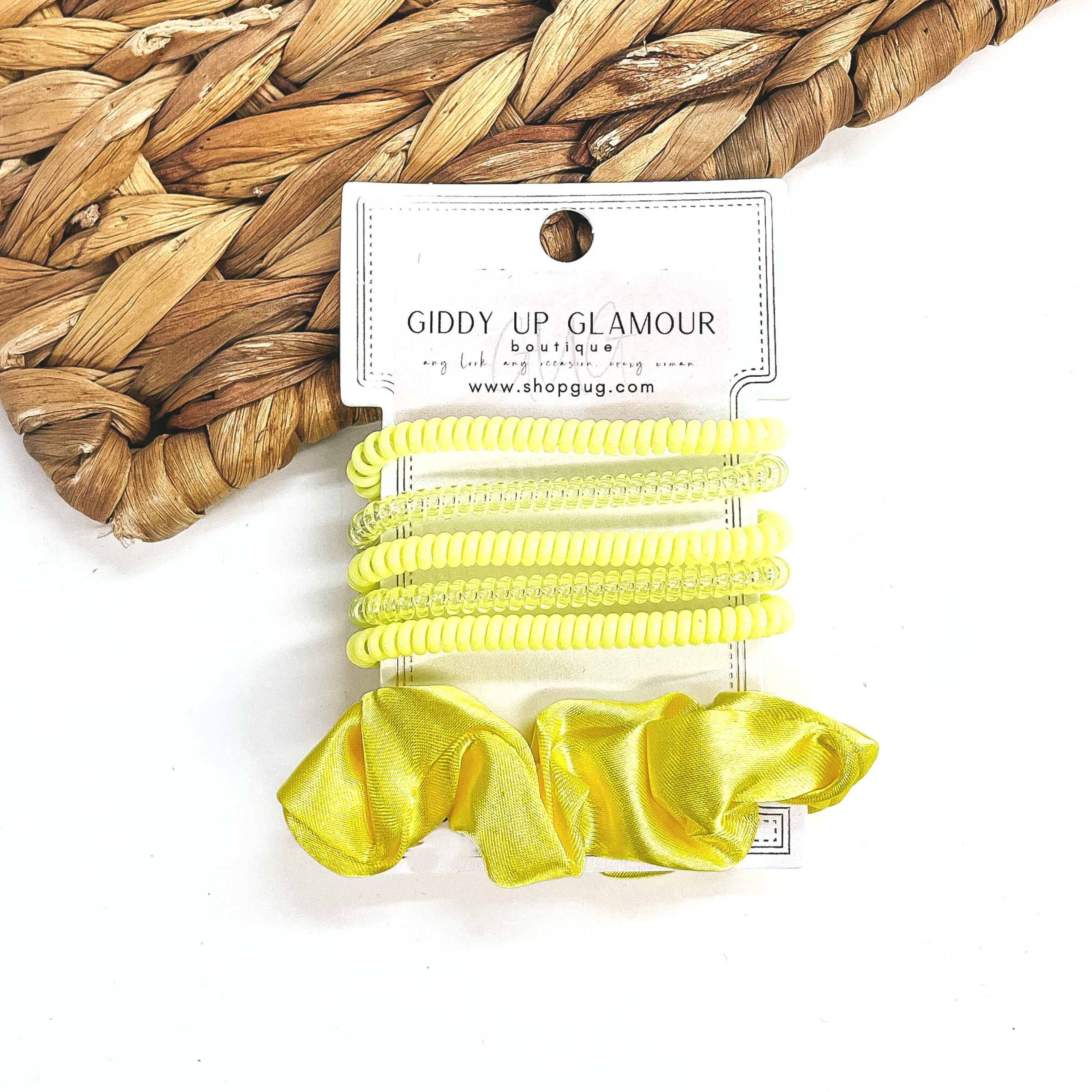 A set of six hair ties, five spiral and one satin scrunchie in yellow. There are three  solid color spiral hair ties and two transparent. These hair ties are on a white  cardboard piece with a Giddy Up Glamour logo, they are leaned up against a brown woven  plate and a white background.