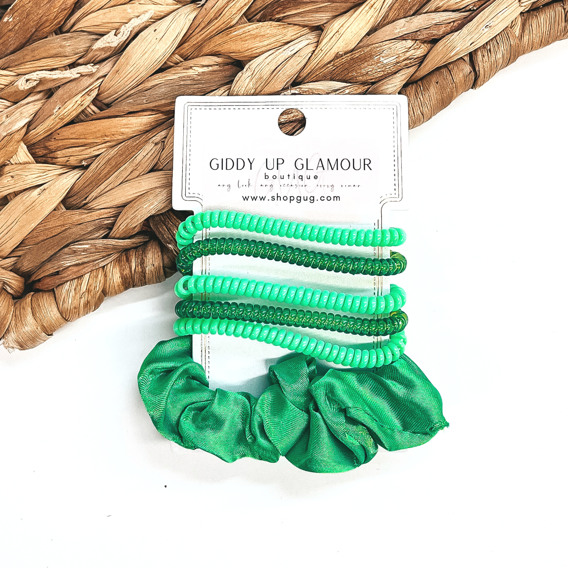 A set of six hair ties, five spiral and one satin scrunchie in green. There are three  solid color spiral hair ties and two transparent. These hair ties are on a white  cardboard piece with a Giddy Up Glamour logo, they are leaned up against a brown woven  plate and a white background.