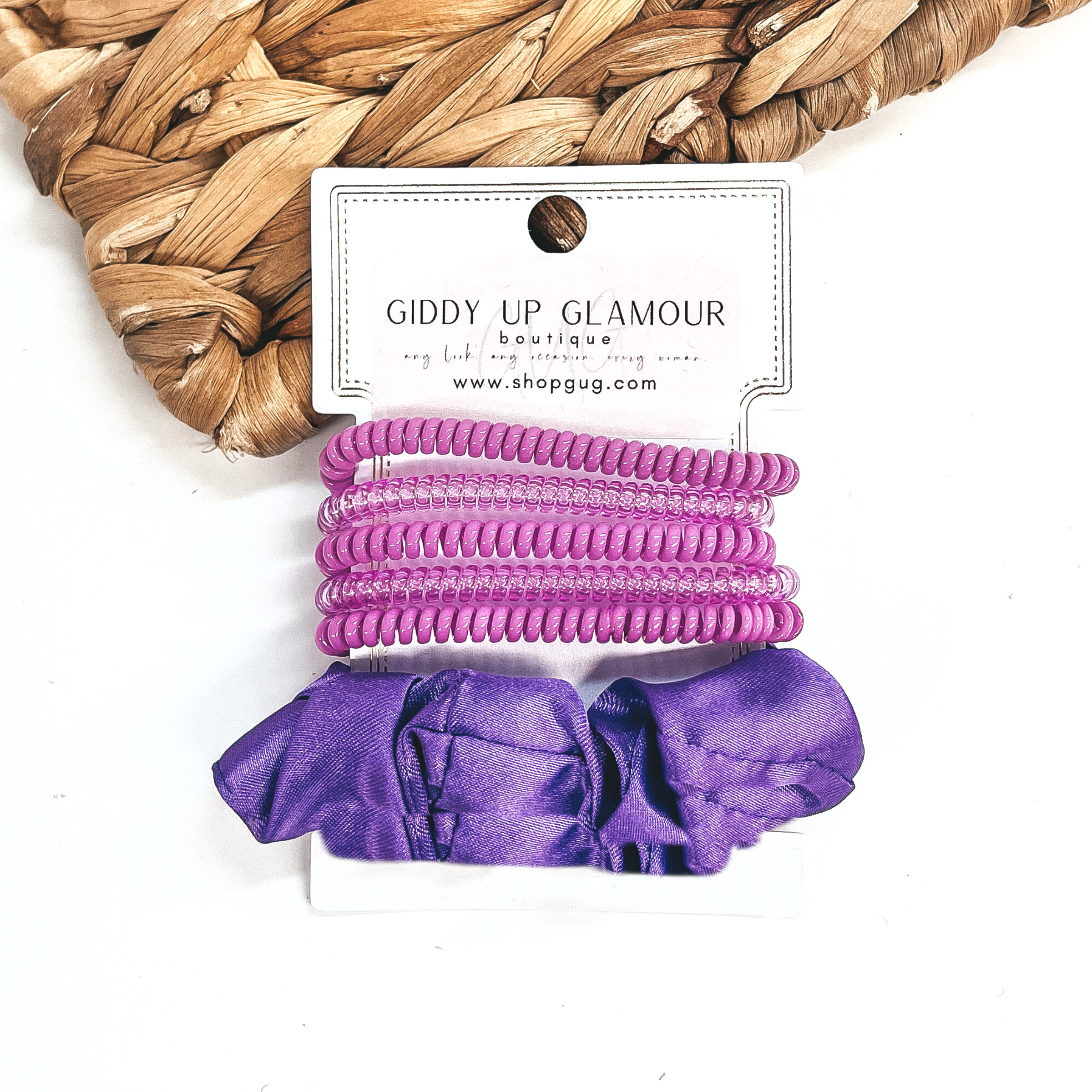 A set of six hair ties, five spiral and one satin scrunchie in purple. There are three  solid color spiral hair ties and two transparent. These hair ties are on a white  cardboard piece with a Giddy Up Glamour logo, they are leaned up against a brown woven  plate and a white background.