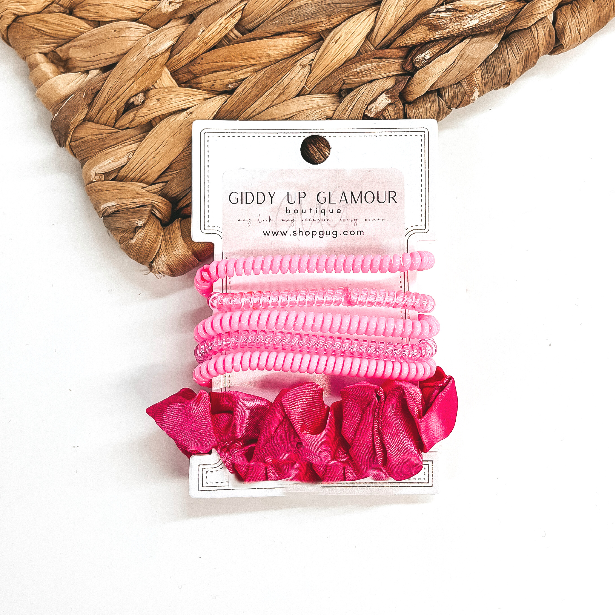 A set of six hair ties, five spiral and one satin scrunchie in hot pink. There are three  solid color spiral hair ties and two transparent. These hair ties are on a white  cardboard piece with a Giddy Up Glamour logo, they are leaned up against a brown woven  plate and a white background.