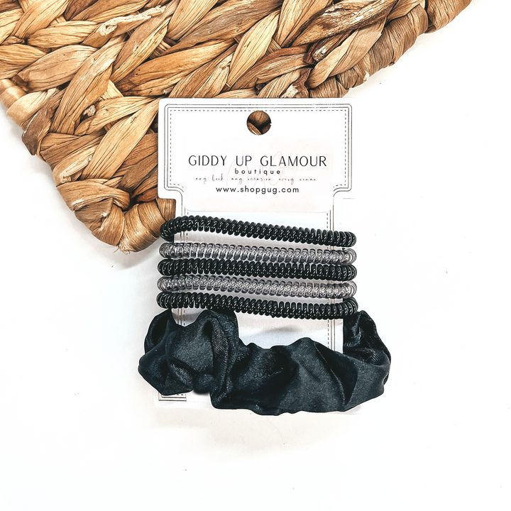 A set of six hair ties, five spiral and one satin scrunchie in black. There are three  solid color spiral hair ties and two transparent. These hair ties are on a white  cardboard piece with a Giddy Up Glamour logo, they are leaned up against a brown woven  plate and a white background.