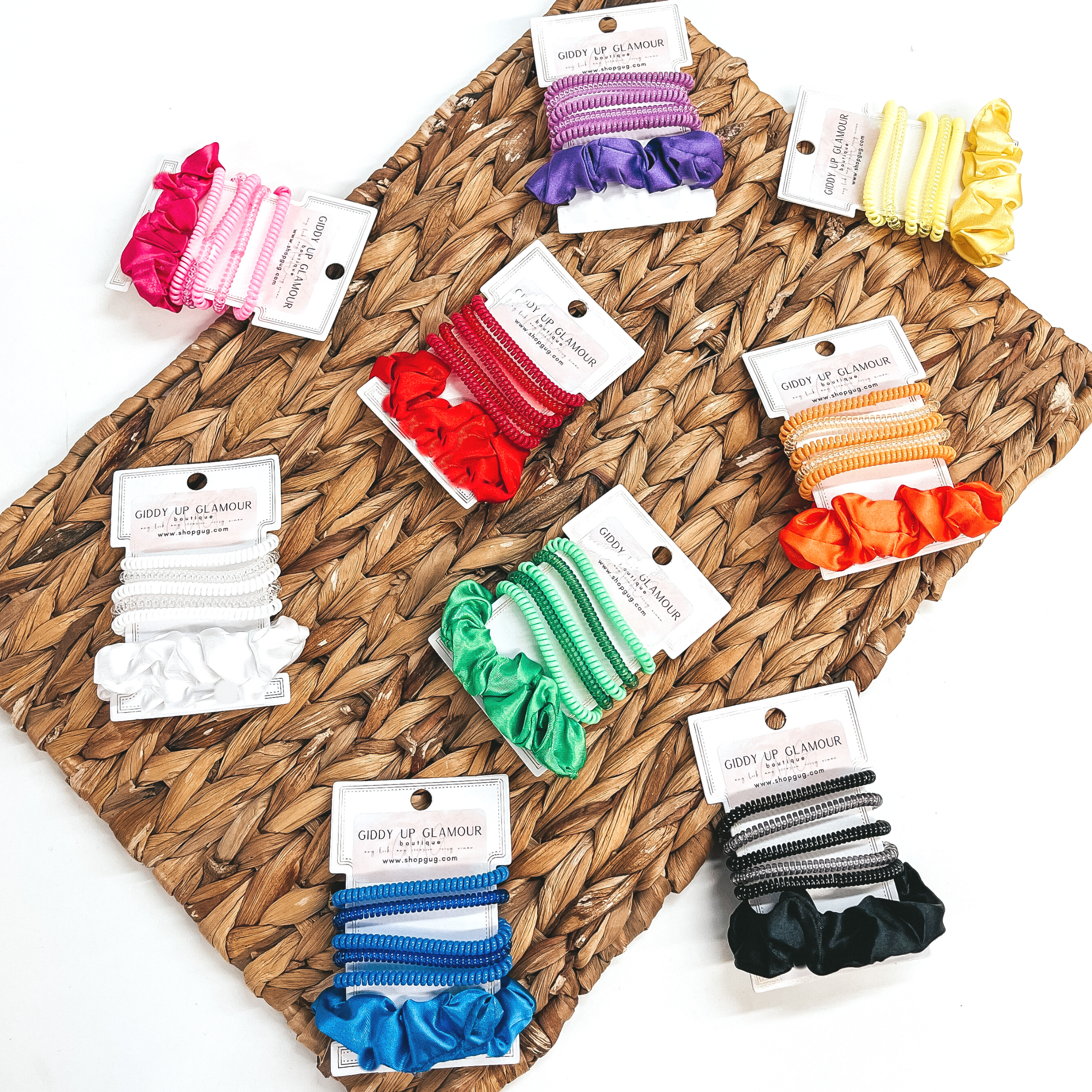 There are nine sets of six spiral and scrunchie hair ties in different colors.  Each set has five spiral hair ties, two transparent and three solid, with one solid  color satin scrunchie. There is in purple, hot pink, yellow, green, orange, red, white, black, and blue. These sets of scrunchies are pictured on a white hair tie  holder. They are laying on a brown woven plate and on a white background.