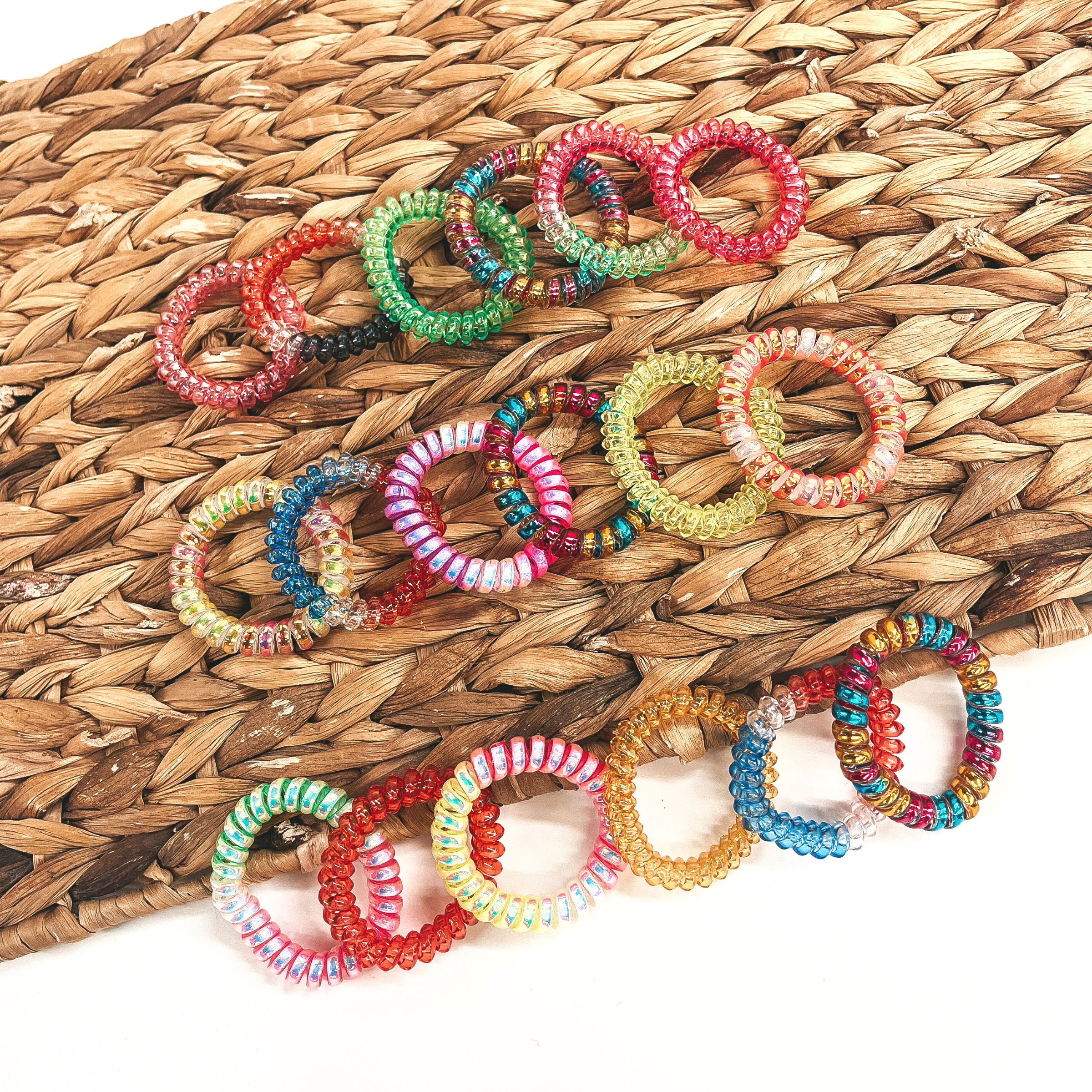 There are fifteen multicolor spiral scrunchies laying on a brown woven plate and on  a white background. There are orange, red, pink, yellow, green solid colors. Also  some in ombre with clear, red, and blue and others with sparkles. 