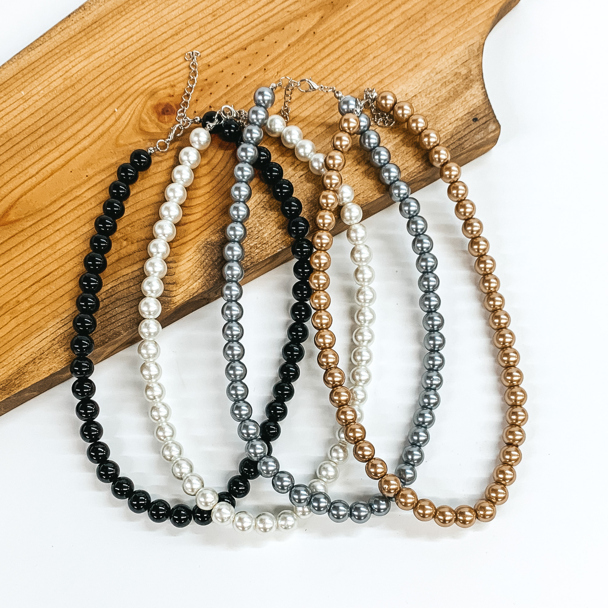 Pictured are four pearl beaded necklaces partially laying on a wood block on a white background. These necklaces are in the colors black, ivory, grey, and bronze.