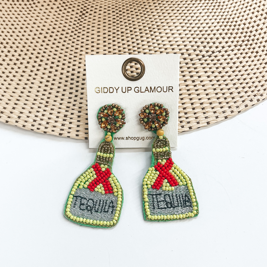 Bronze crystal and brown beaded studs with hanging beaded bottle. The bottle has a gold beaded top with a lime green beaded body with red beaded ribbon. The center on the bottom is grey stitched with the word "TEQUILA" stitched in black. These earrings are pictured on a white earrings holder laying partially on a straw hat brim on a white background.
