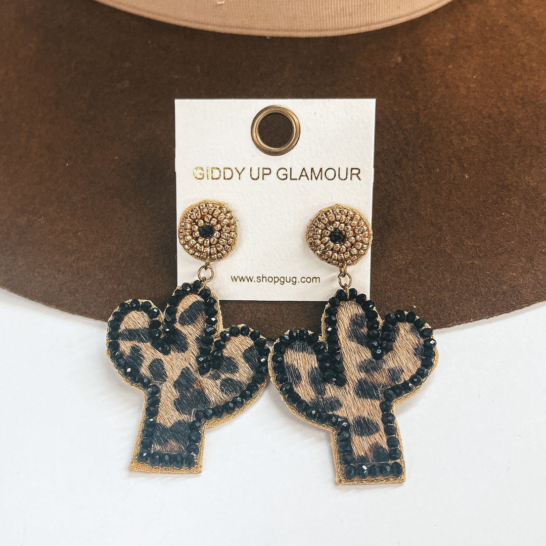 Cactus shape earrings with leopard print, black and gold beads.  The leopard print is in faux hide and has black beads all around as  the outline. The post back part has gold beads and one black bead in  the center. These earrings are taken on a dark brown hat and on a white  background.