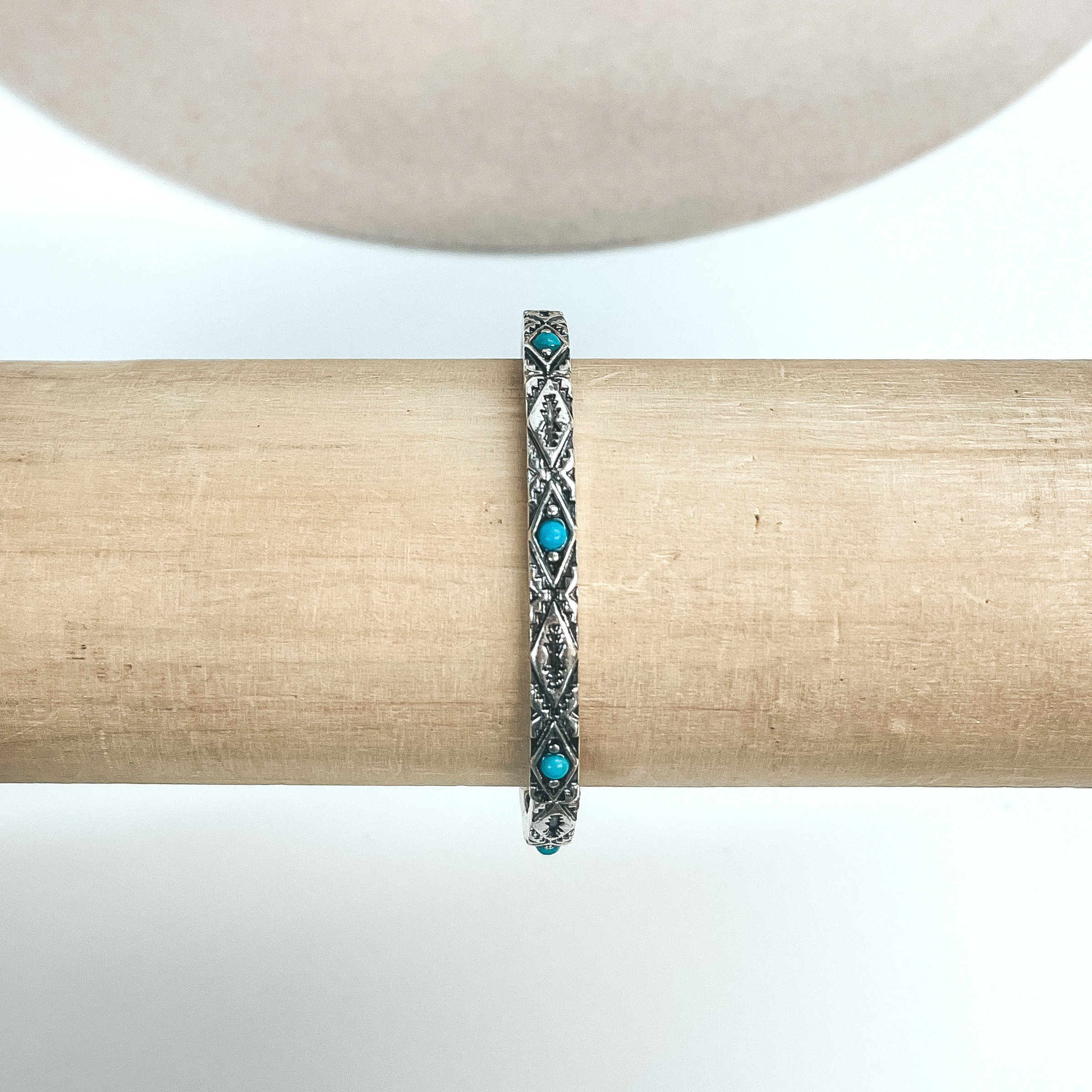 This is a thin silver bracelet with Aztec engraved design and small  turquoise stones all around in the center. These earrings are taken on a  light brown bracelet holder and on  a white background with a beige hat in the back as decor.