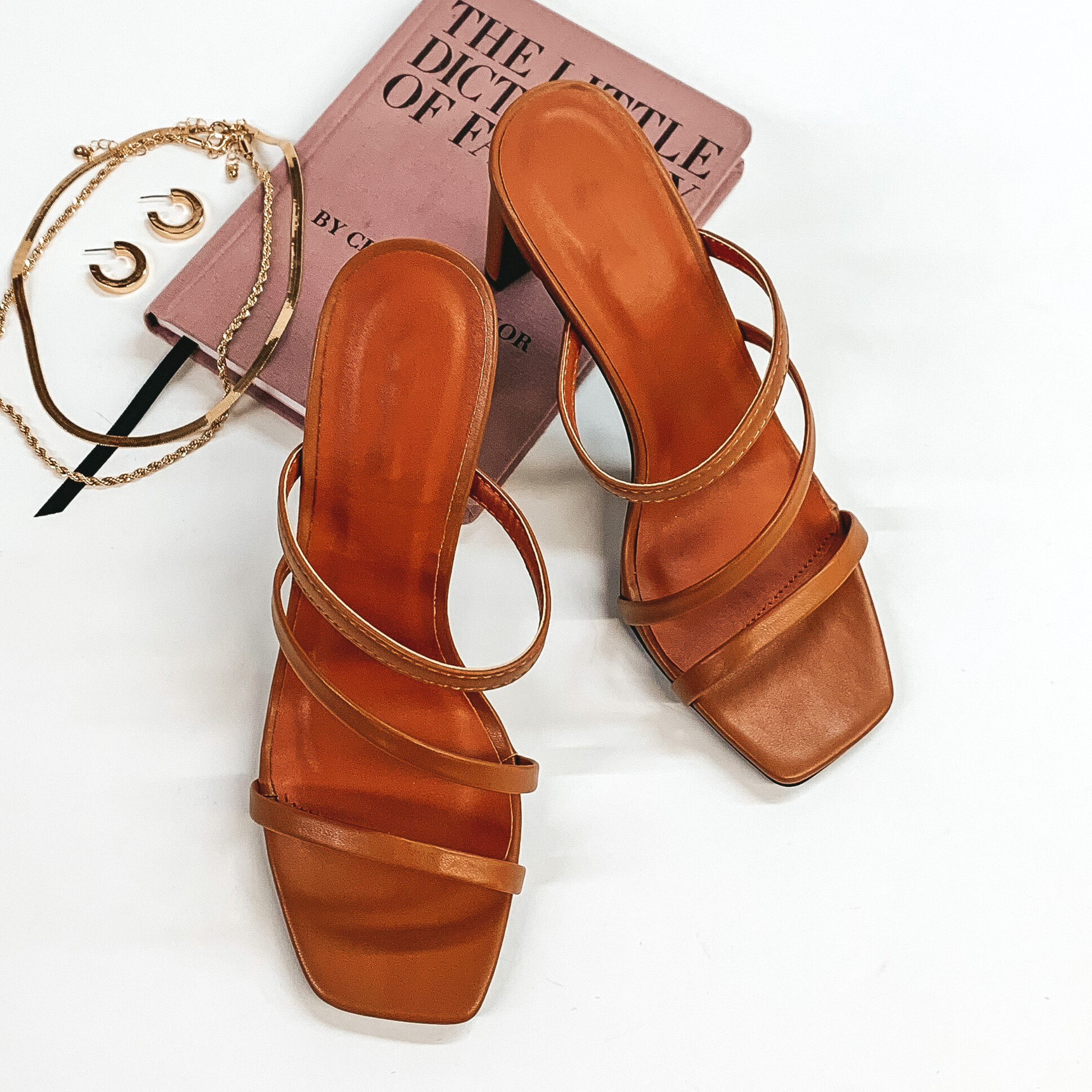 A pair of camel colored strappy heels that have a square toe. These shoes are pictured on a white background with gold jewelry and a book.