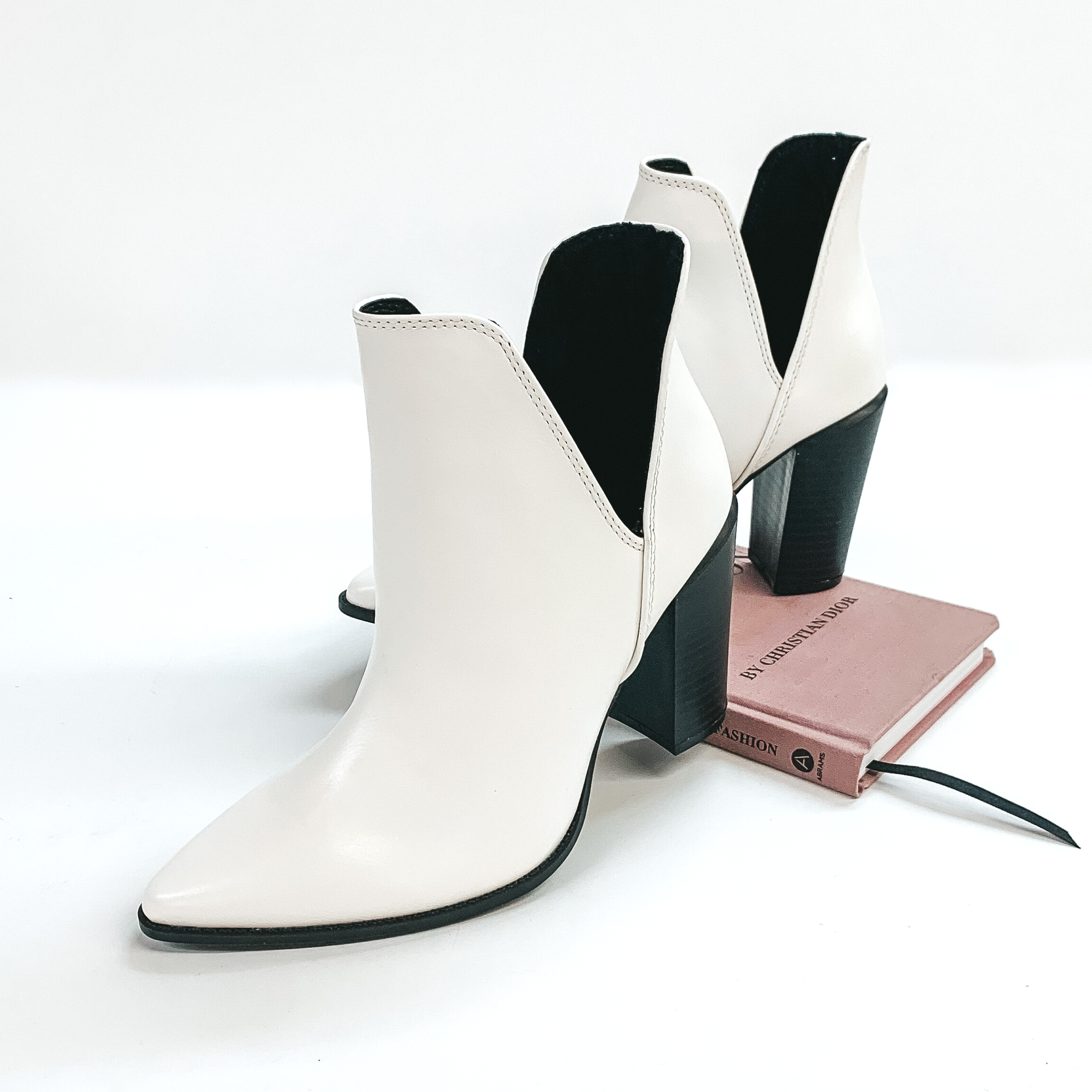 Ankle booties in white with a black wooden heel. These booties are pictured on a white background standing on a book. 
