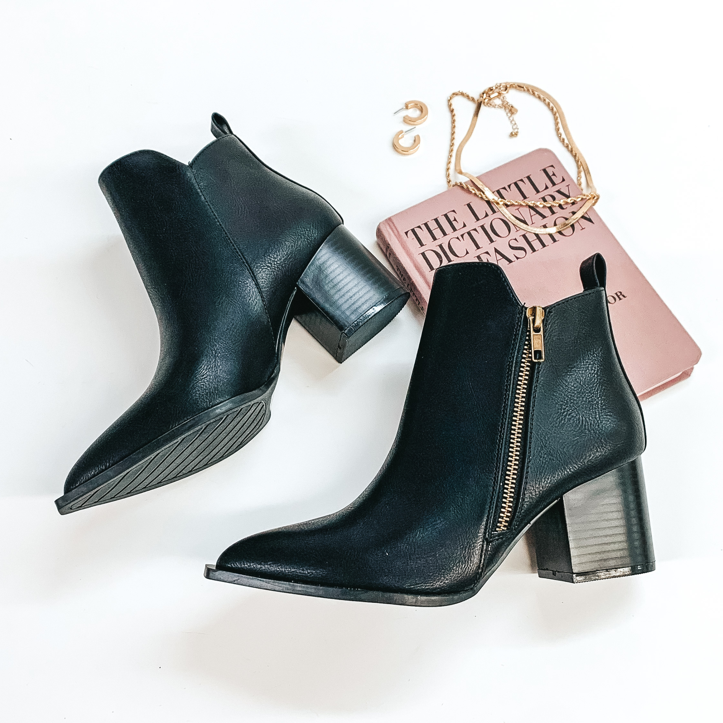 Ankle booties in black with a side zipper. These booties also have a wooden heel. These booties are pictured on a white background with gold jewelry and a book.  
