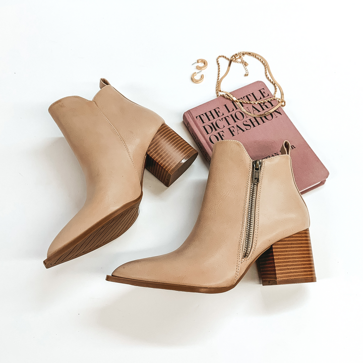 Ankle booties in beige with a side zipper. These booties also have a wooden heel. These booties are pictured on a white background with gold jewelry and a book.  