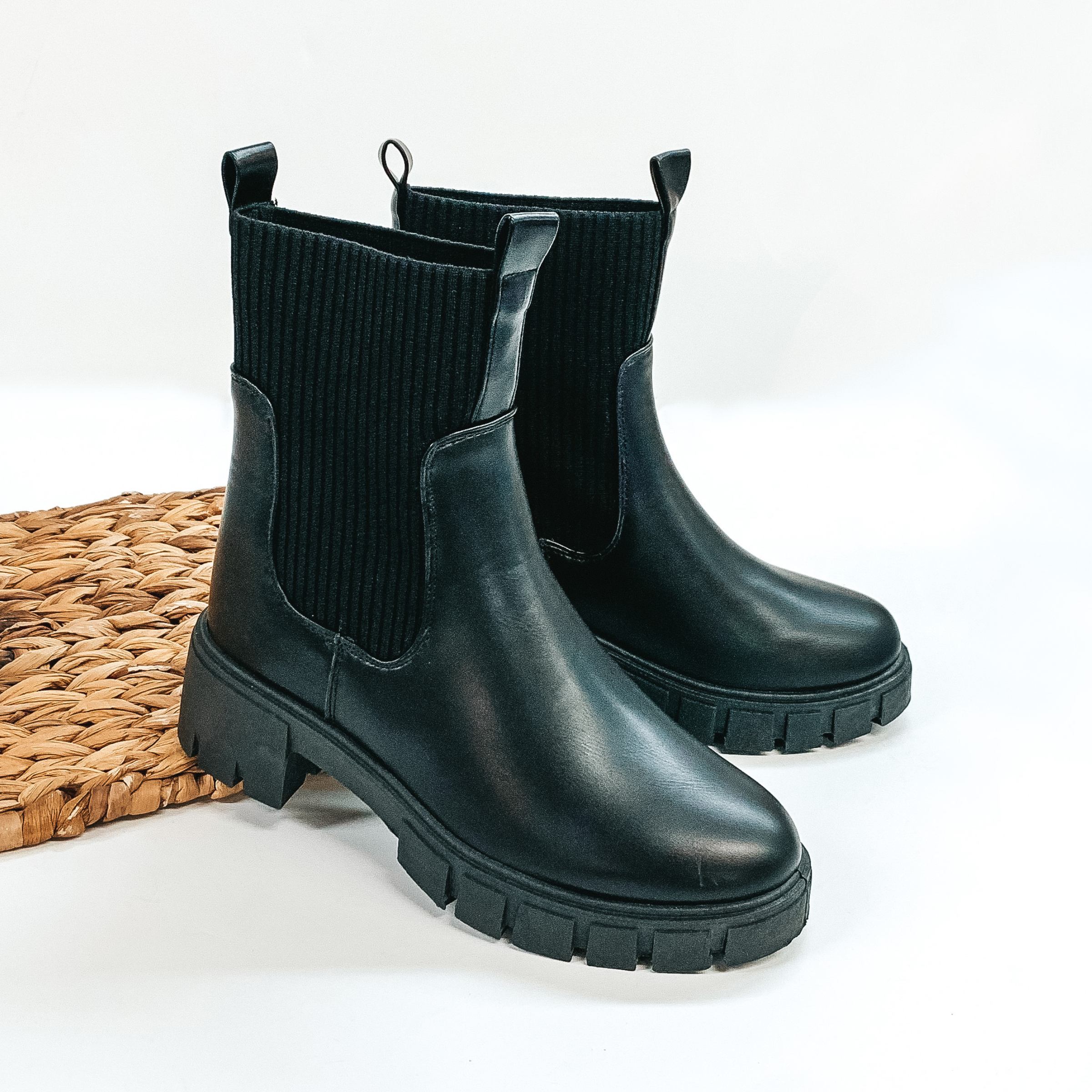 Ankle high black colored slip on bootie. The sides are elastic and also have a front and back pull on tab. These booties are pictured propped on a basket weave material on a white background. 