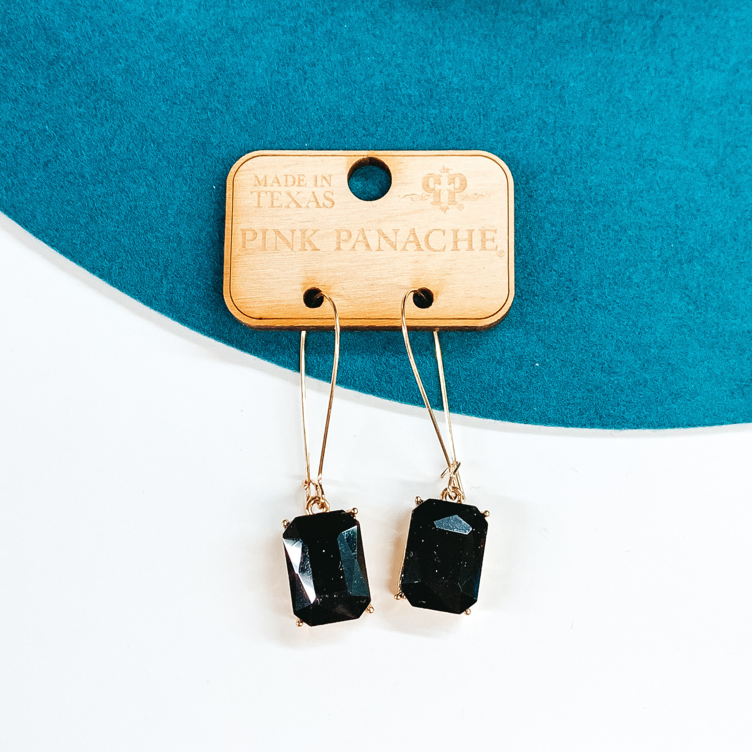 Gold kidney wire earrings with a hanging rectangle black crystal pendant. These earrings are pictured on a teal and white background. 
