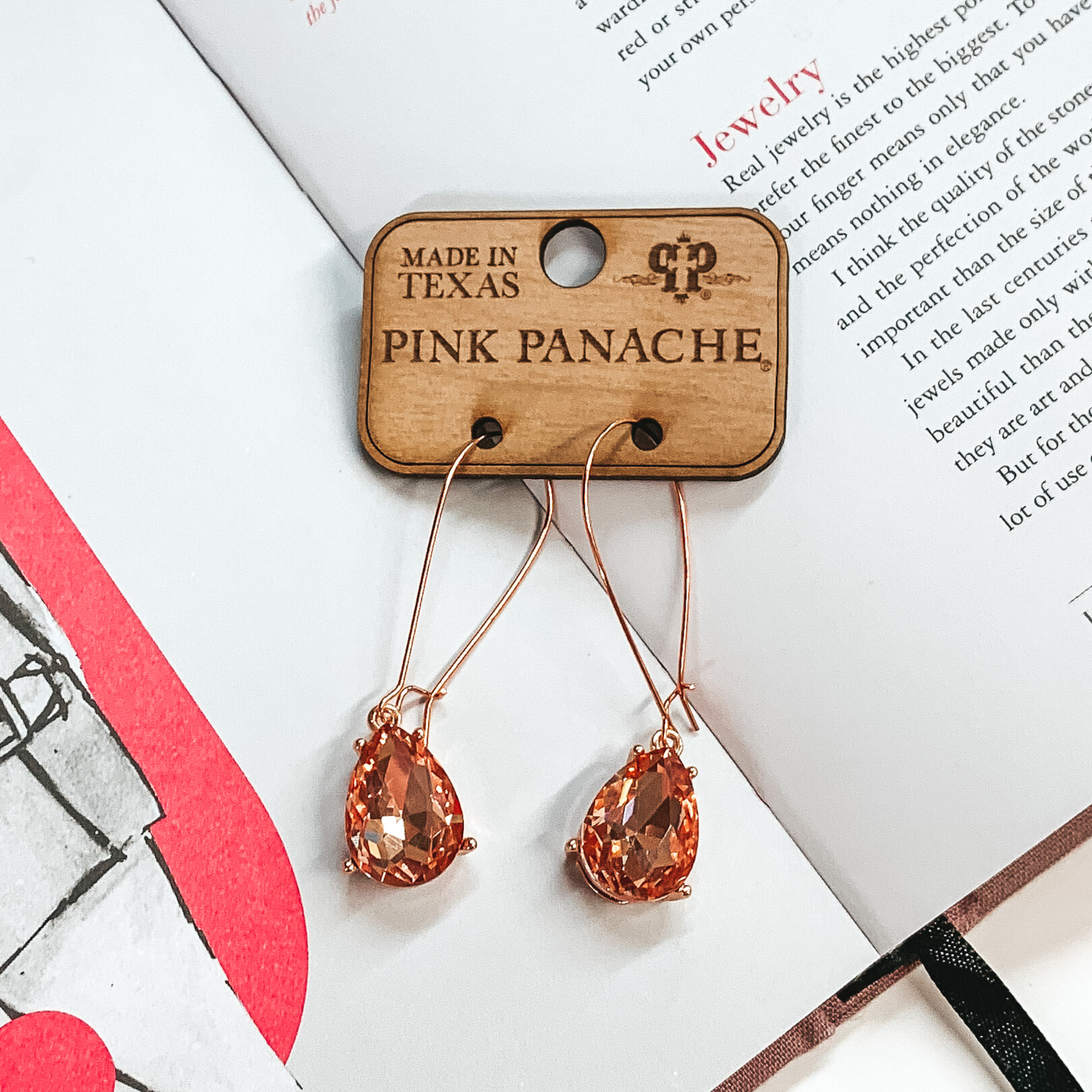 Rose gold kidney wire earrings with a hanging teardrop rose gold crystal pendant. These earrings are pictured on an open book.