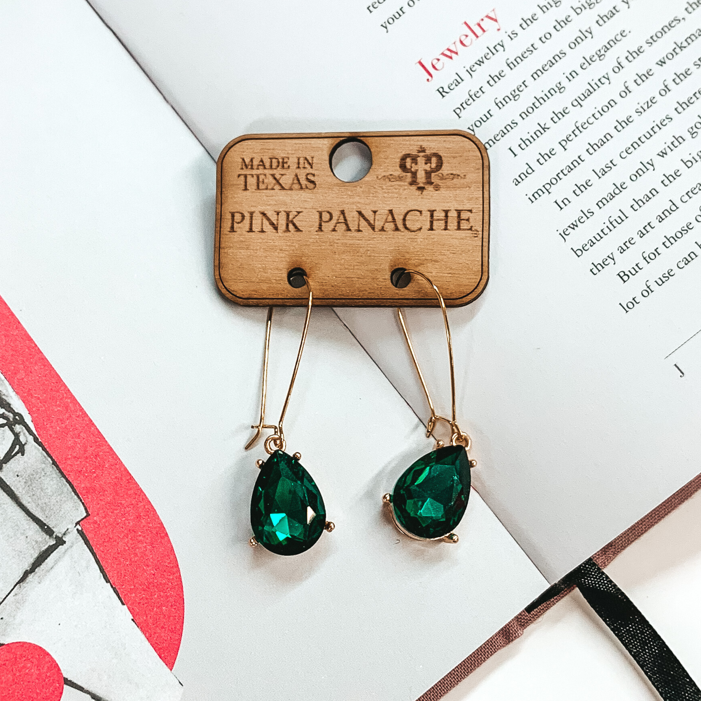 Gold kidney wire earrings with a hanging teardrop emerald crystal pendant. These earrings are pictured on an open book.
