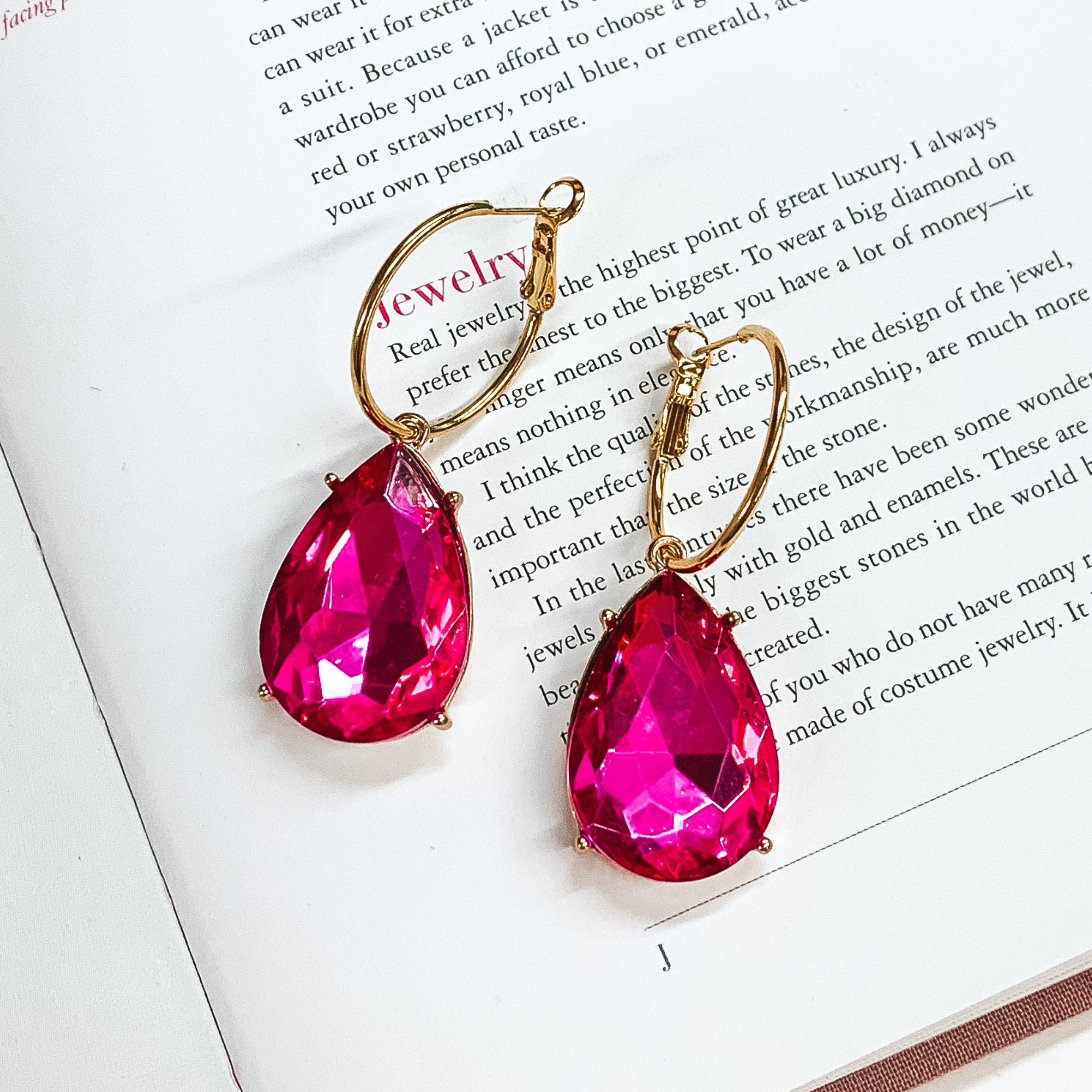 A pair of gold hoop earrings with fuchsia teardrop crystals. These earrings are pictured on an open book.