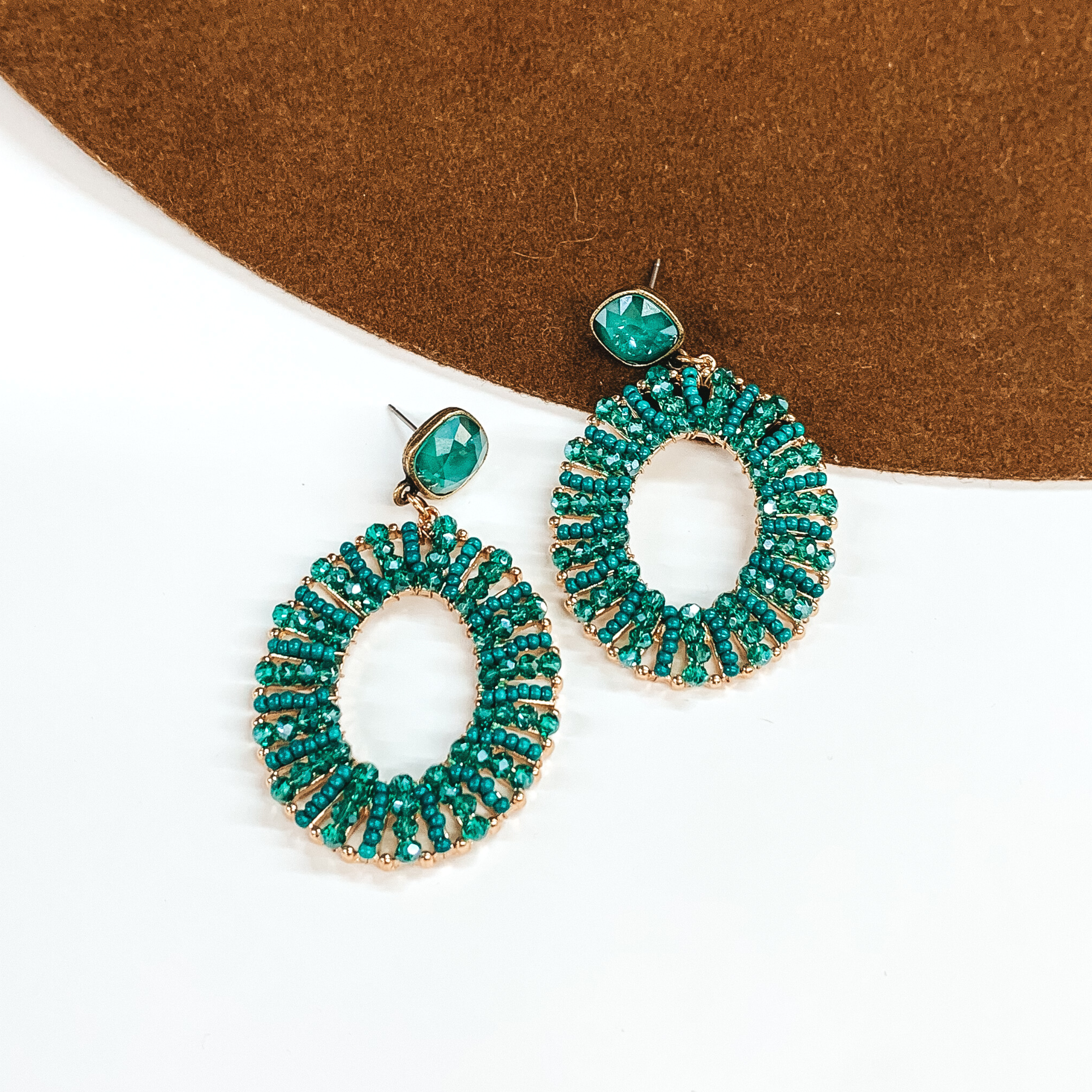 Pair of open oval, beaded earrings. These earrings include royal green cushion cut crystal stud earrings and the oval pendant has green beads. These earrings are pictured laying partially on a brown hat brim on a white background.