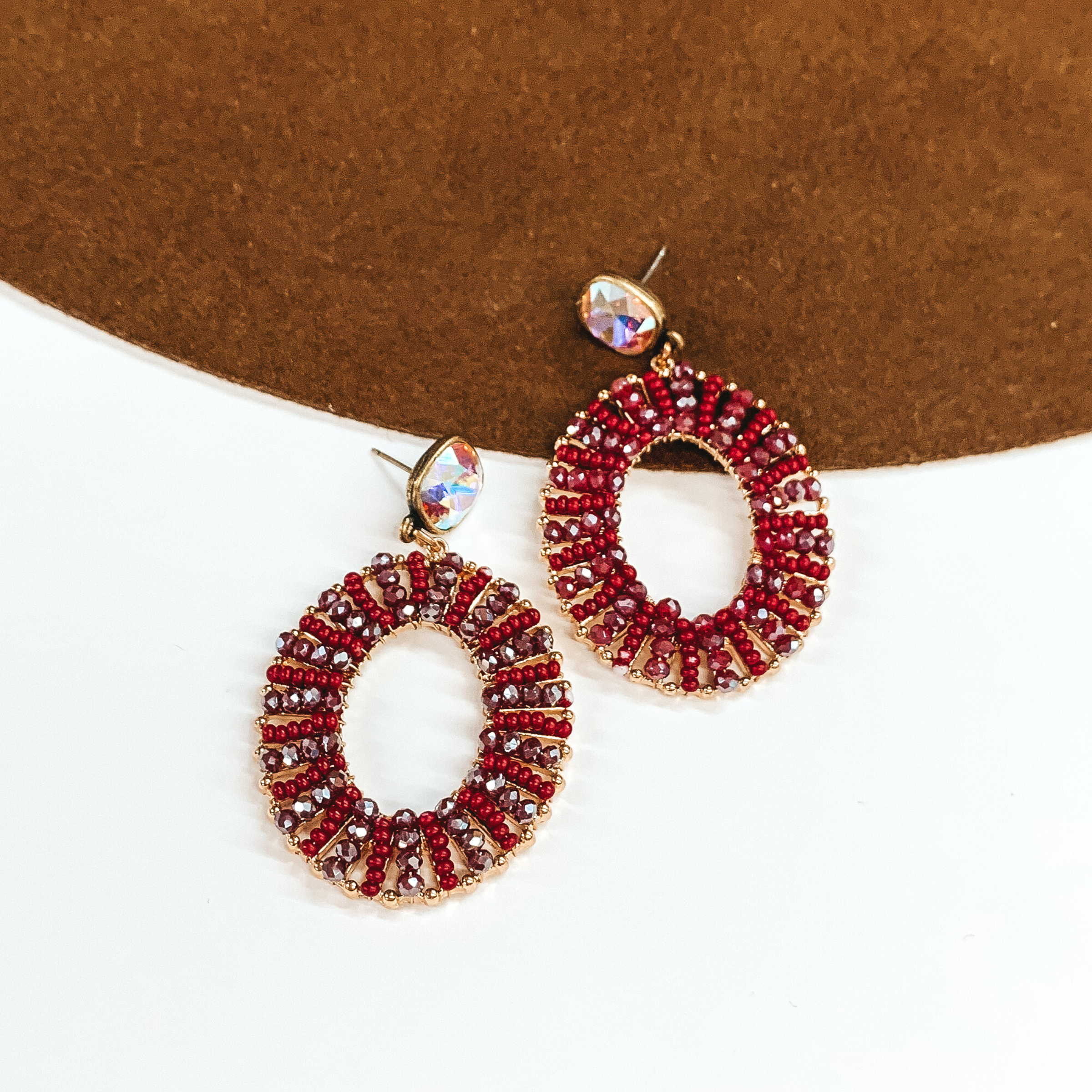 Pair of open oval, beaded earrings. These earrings include ab cushion cut crystal stud earrings and the oval pendant has burgundy beads. These earrings are pictured laying partially on a brown hat brim on a white background. 