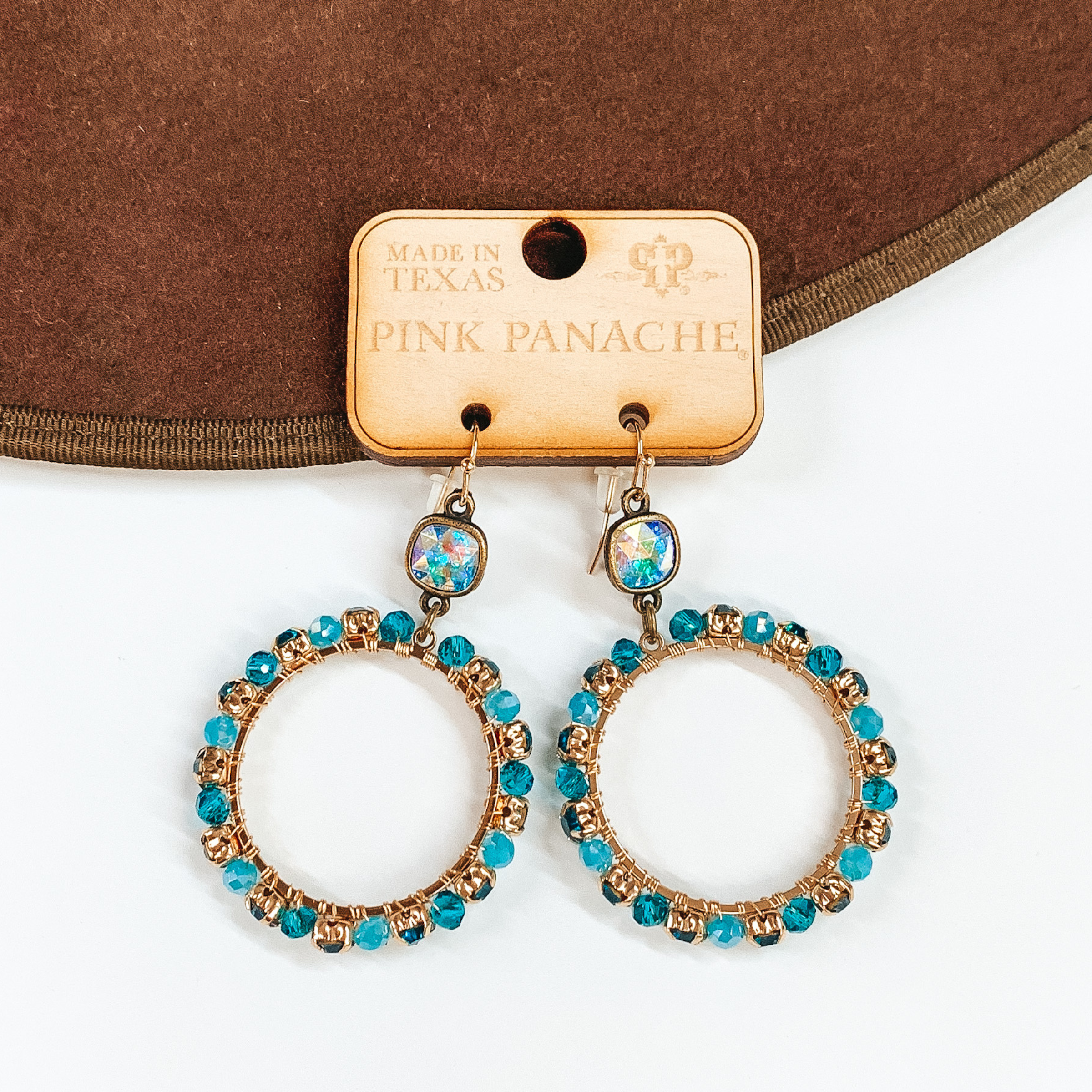 Pictured is a pair of circle hoop earrings. These earrings include gold fish hook earrings, ab cushion cut crystal connectors, and the hoop is gold with dark and light awua colored beads and crystals. These earrings are pictured laying partially on a brown hat brim on a white background.  