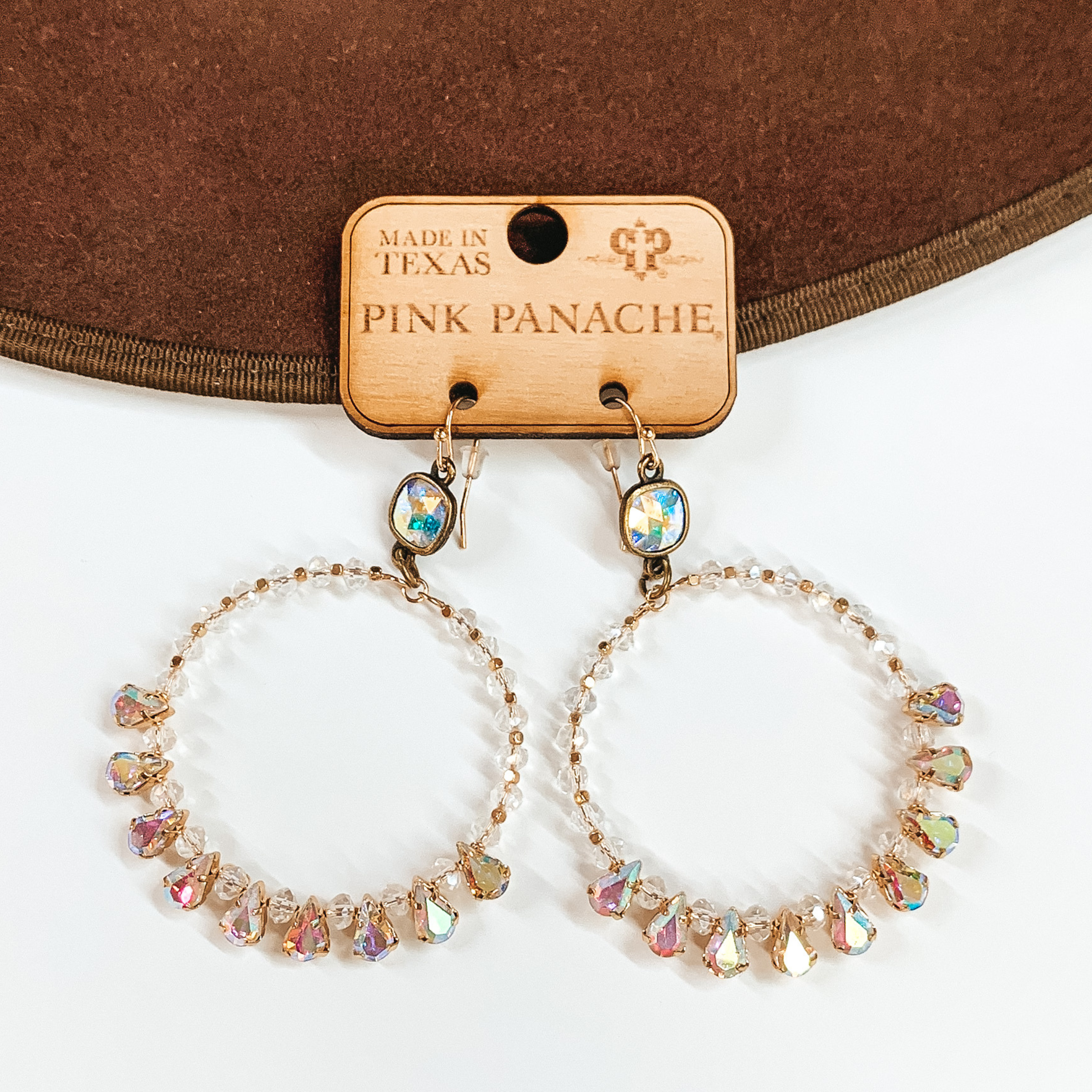 Pictured is a pair of large, circle hoop earrings. These earrings include gold fish hook earrings, ab cushion cut crystal connectors, and the hoop has gold, clear, and ab crystals. These earrings are pictured laying partially on a brown hat brim on a white background.  
