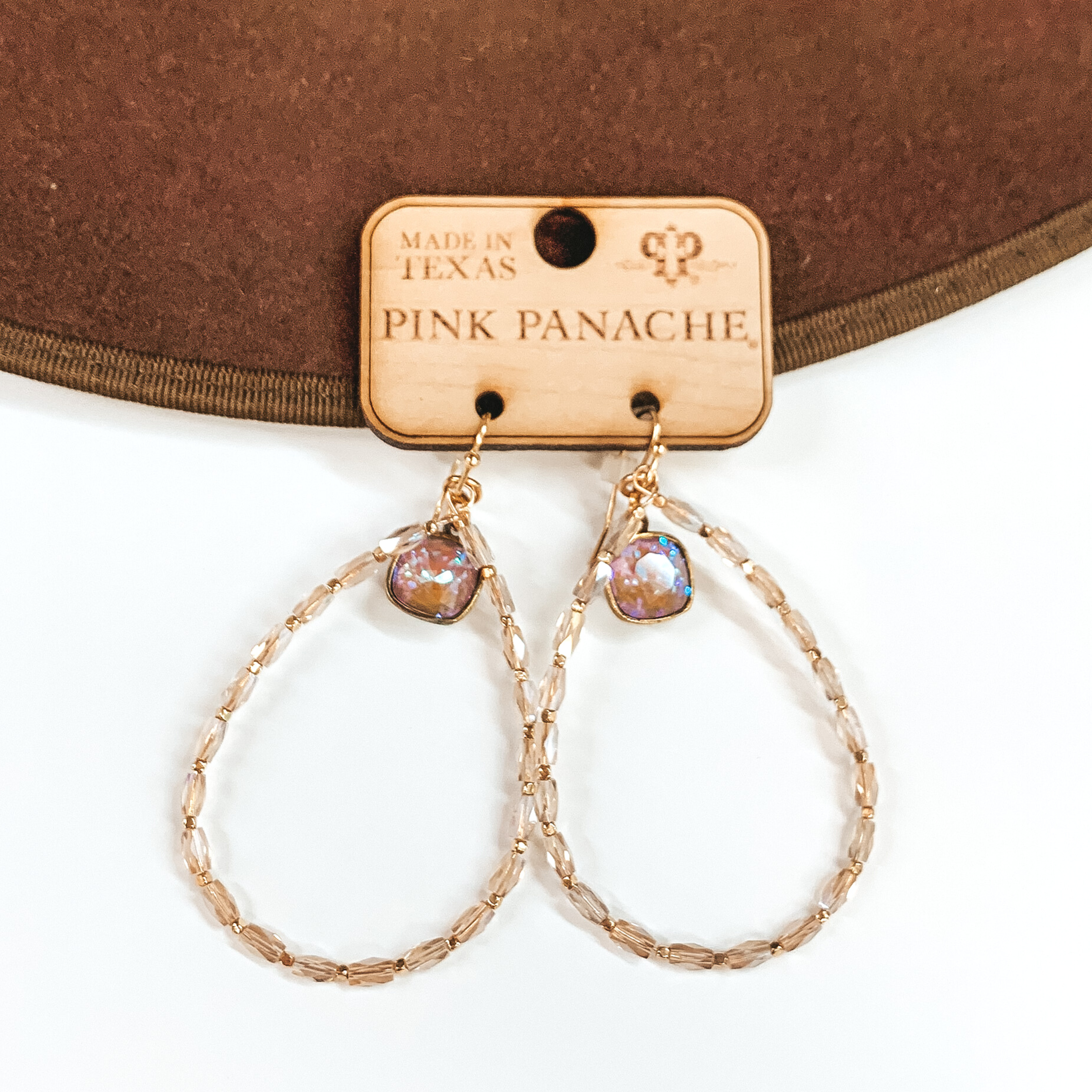 Pictured is a pair of champagne and gold beaded teardrop earrings. These earrings also include cappaccino delight cushion cut drops at the top of the teardrop. These earrings are pictured laying partially on a brown hat brim on a white background.