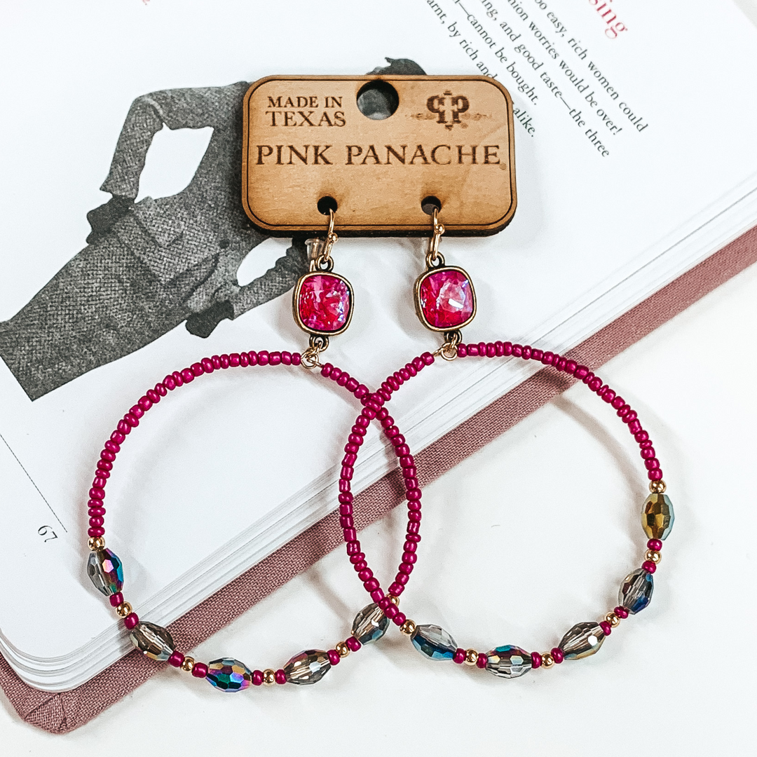 Pictured is a pair of large, circle hoop earrings. These earrings include gold fish hook earrings, royal red delight cushion cut connectors, and the hoop has fuchsia beads and rainbow beads. These earrings are pictured laying on an open book on a white background. 