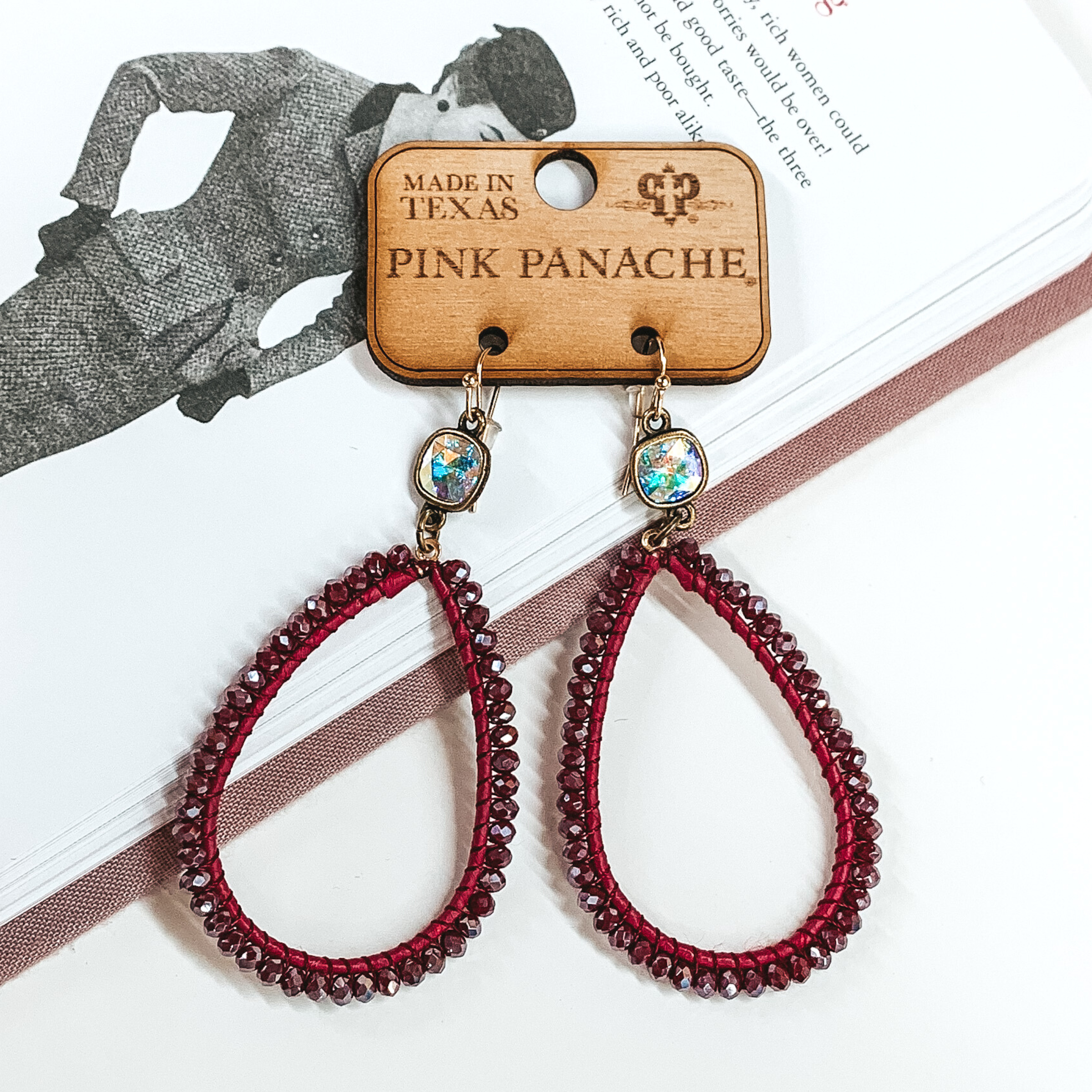 Pair of teardrop earrings. These earrings include gold fish hook earrings, ab cushion cut crystal connectors, and burgundy colored wrap and beads on the teardrop pendant. These earrings are pictured laying on an open book on a white background.