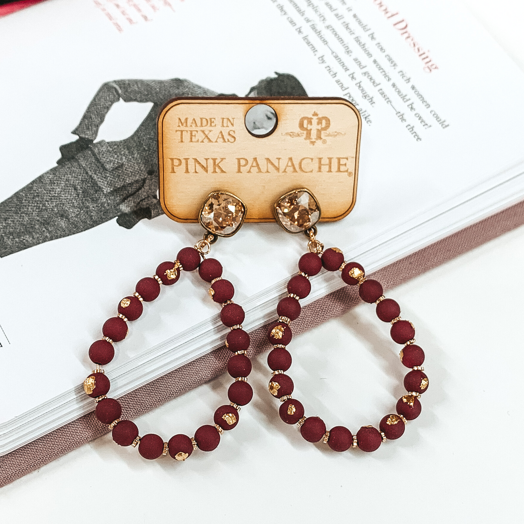 Pair of teardrop earrings. These earrings include golden shadow cushion cut stud earrings with burgundy beaded teardrop pendant with gold flakes. These earrings are pictured laying on an open book on a white background. 