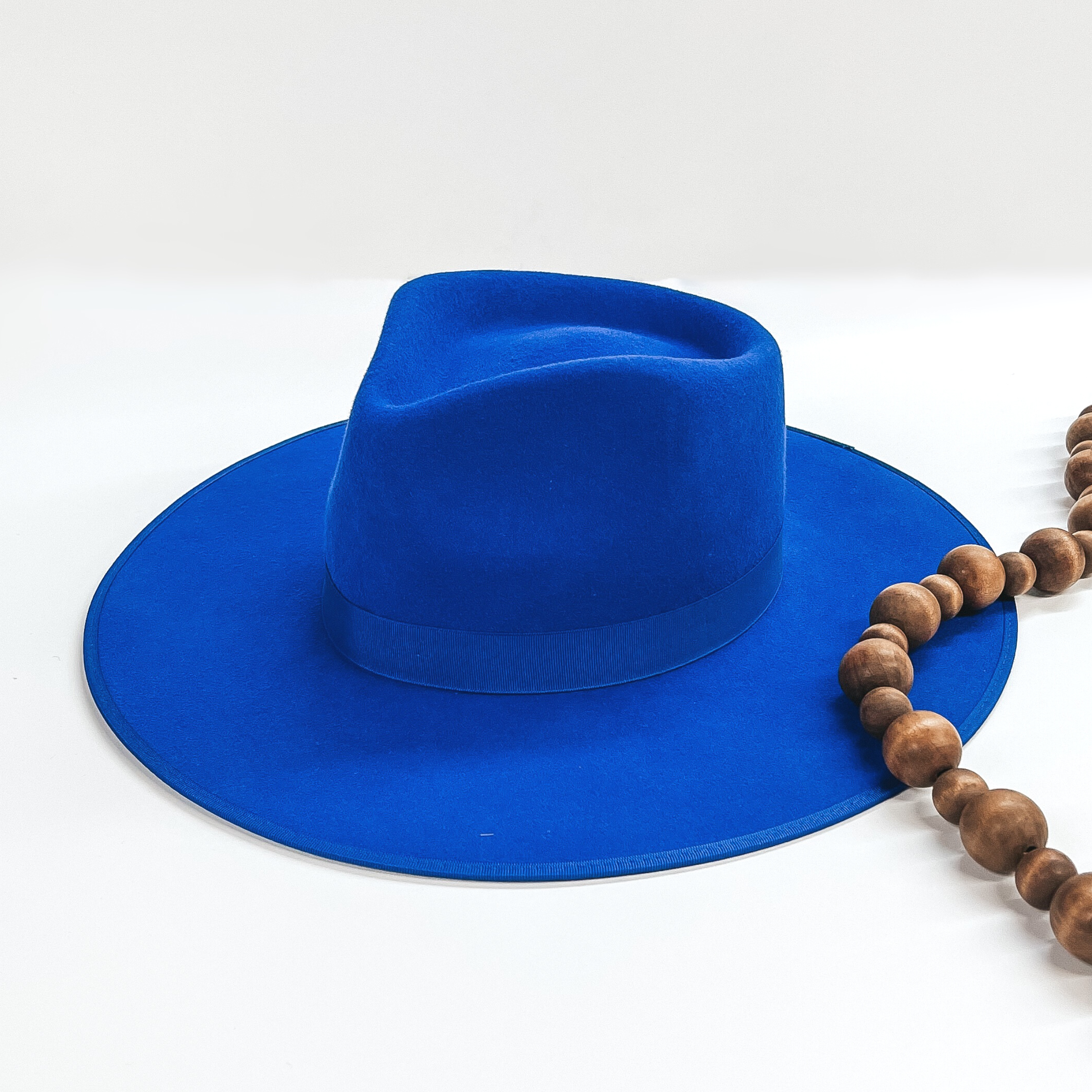 Royal blue felt hat with a structured top and a flat brim. This has includes a royal blue ribbon hat band. This hat is pictured on a white background with brown decorative beads.