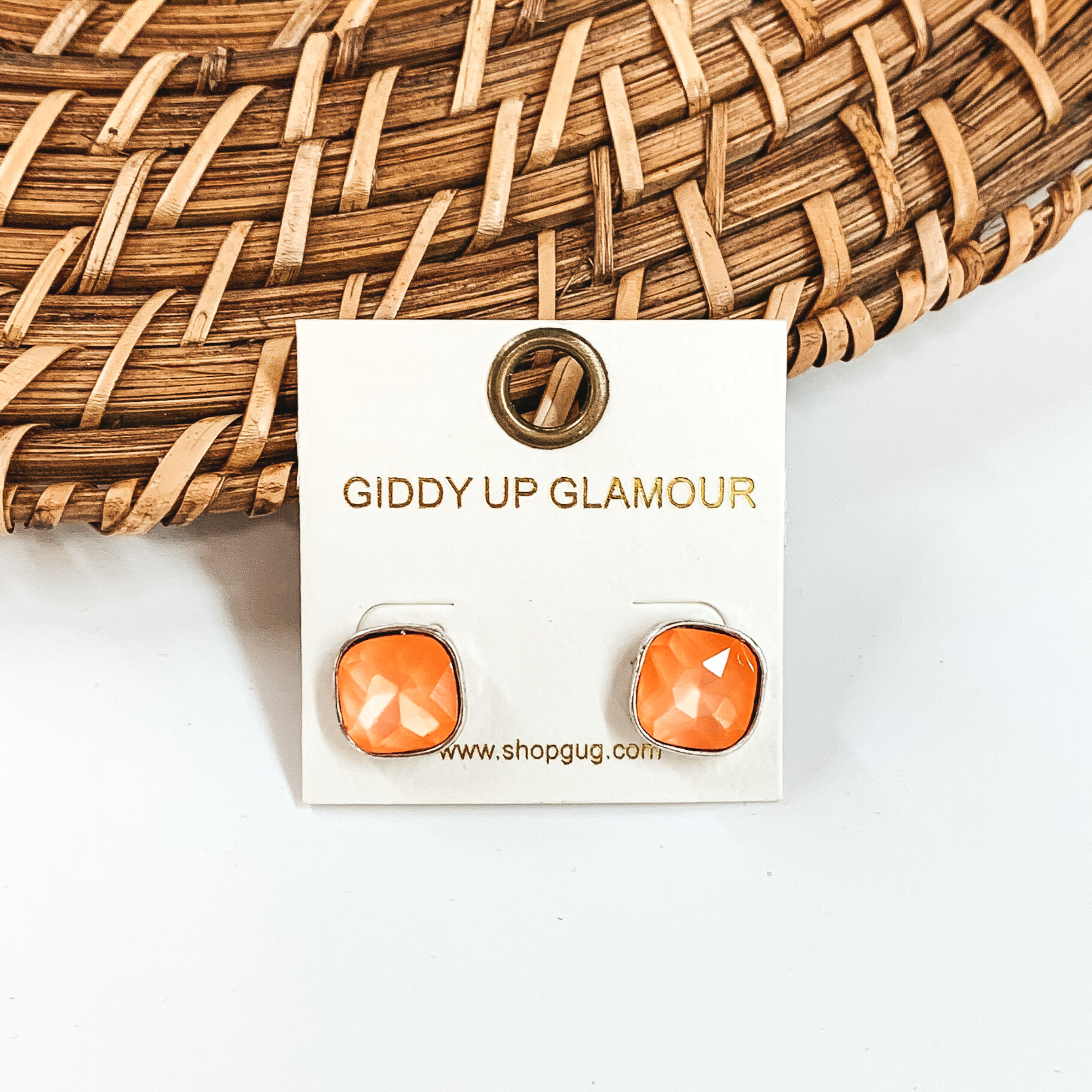 Silver, square stud earrings with orange colored crystals. These earrings are pictured on a white GUG earrings holder on a white background with brown basket weave material behind the earrings. 