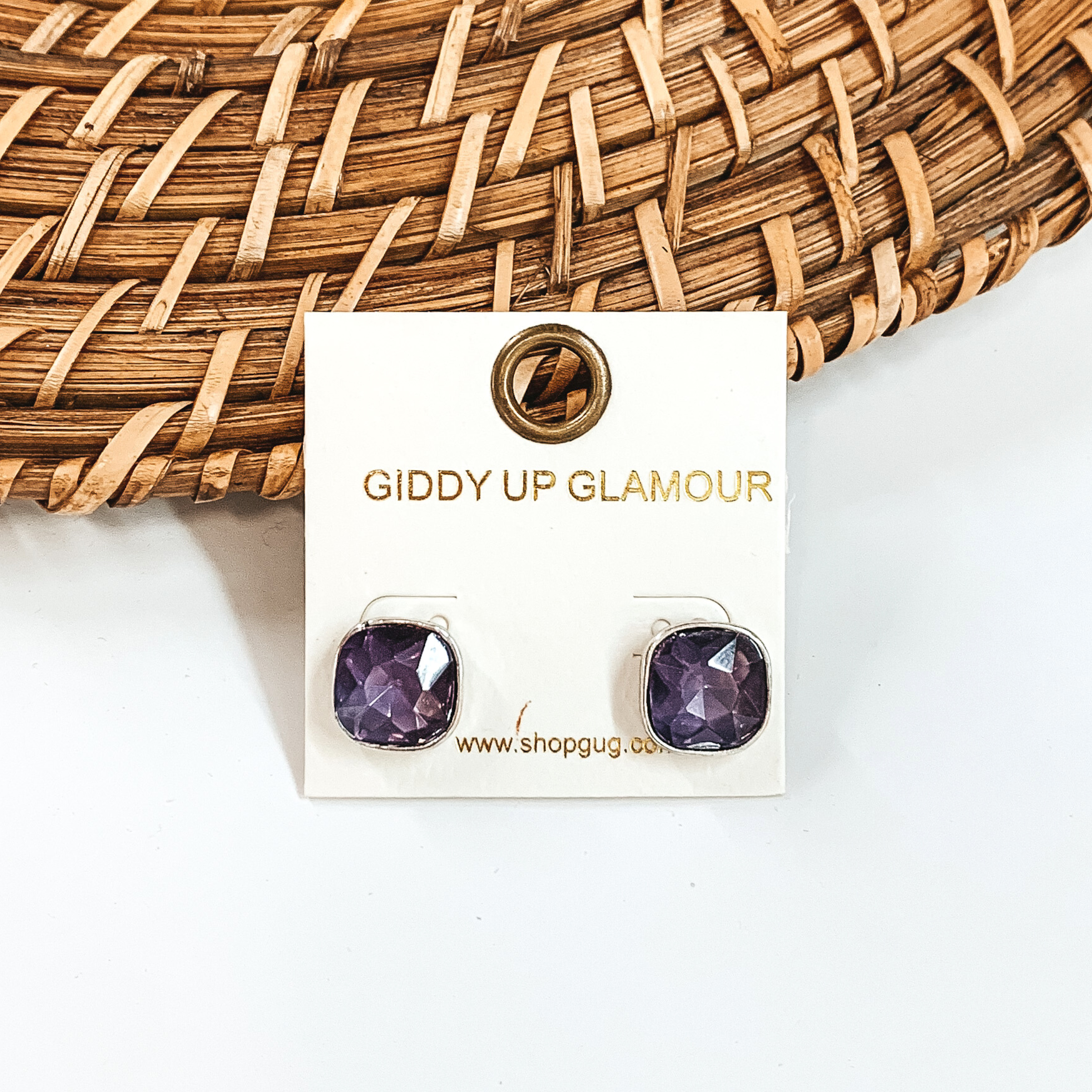Silver, square stud earrings with purple colored crystals. These earrings are pictured on a white GUG earrings holder on a white background with brown basket weave material behind the earrings. 