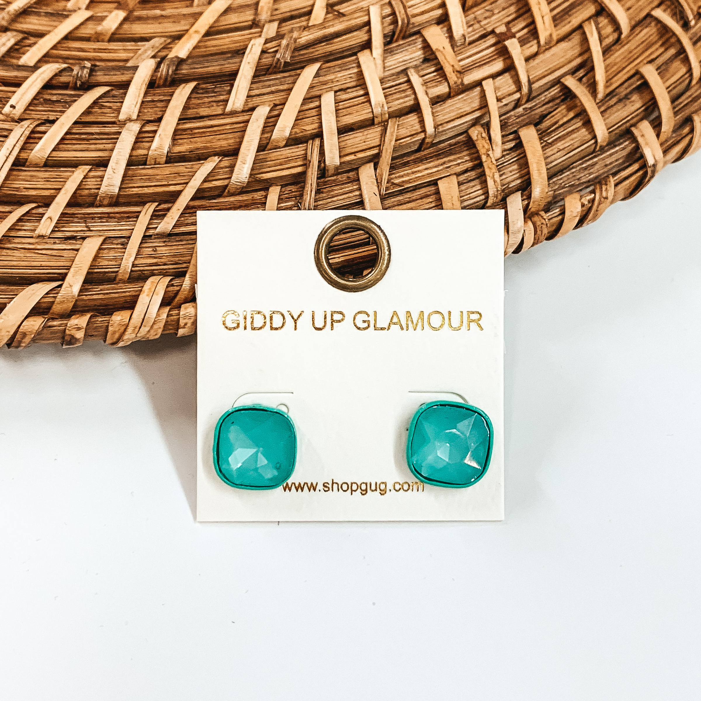 Turquoise, square stud earrings with turquoise colored crystals. These earrings are pictured on a white GUG earrings holder on a white background with brown basket weave material behind the earrings. 