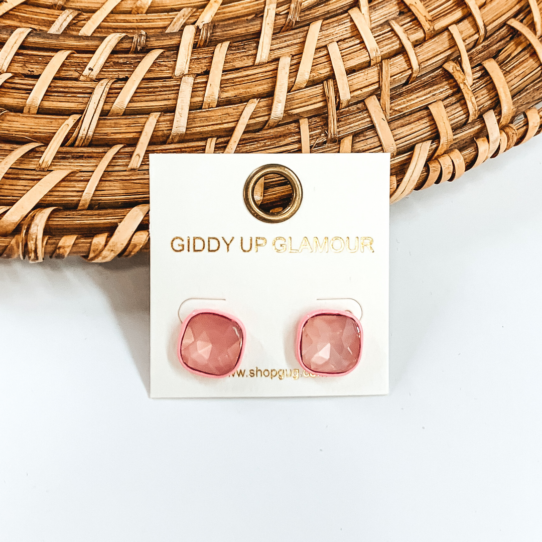 Light pink, square stud earrings with light pink colored crystals. These earrings are pictured on a white GUG earrings holder on a white background with brown basket weave material behind the earrings. 