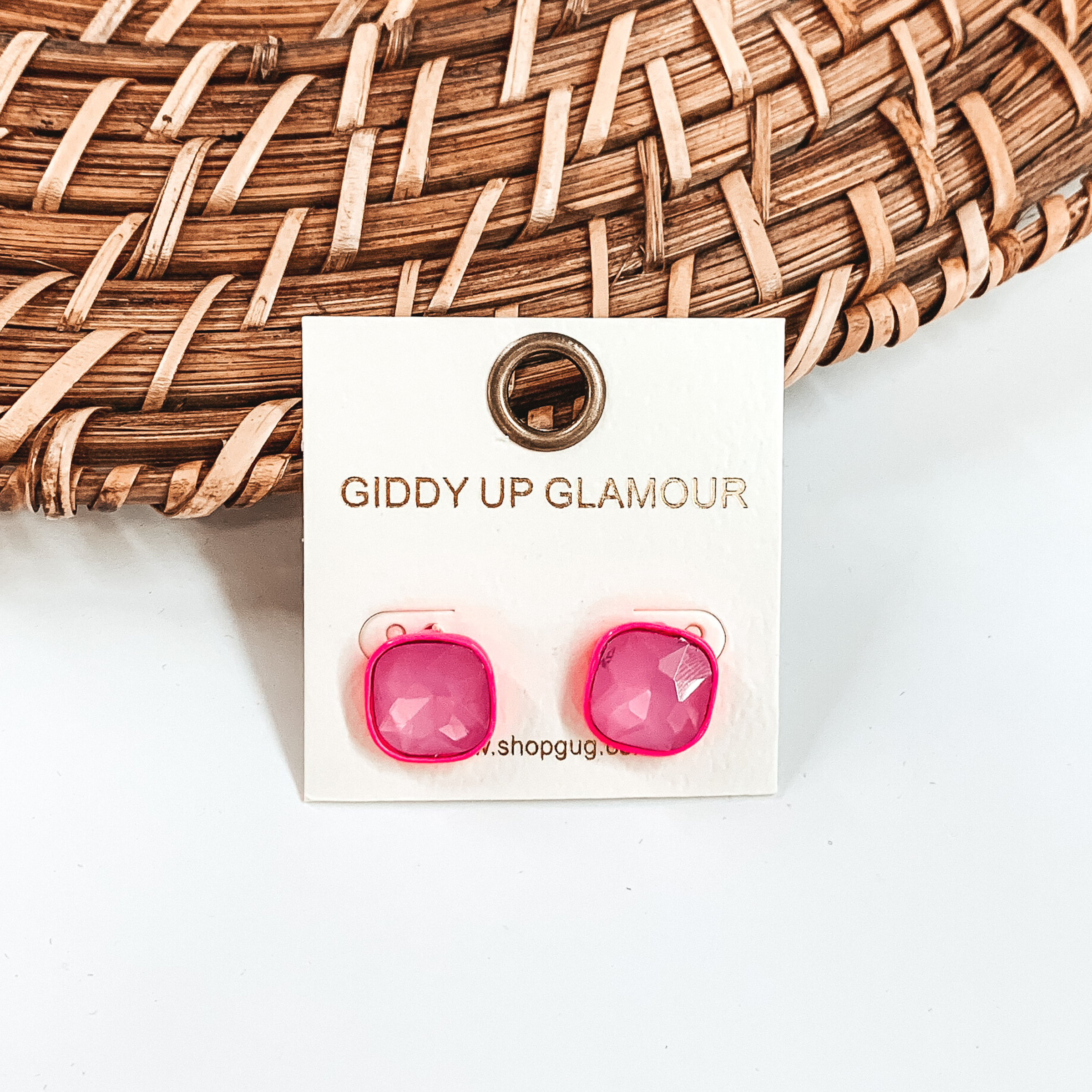 Hot pink, square stud earrings with hot pink colored crystals. These earrings are pictured on a white GUG earrings holder on a white background with brown basket weave material behind the earrings. 