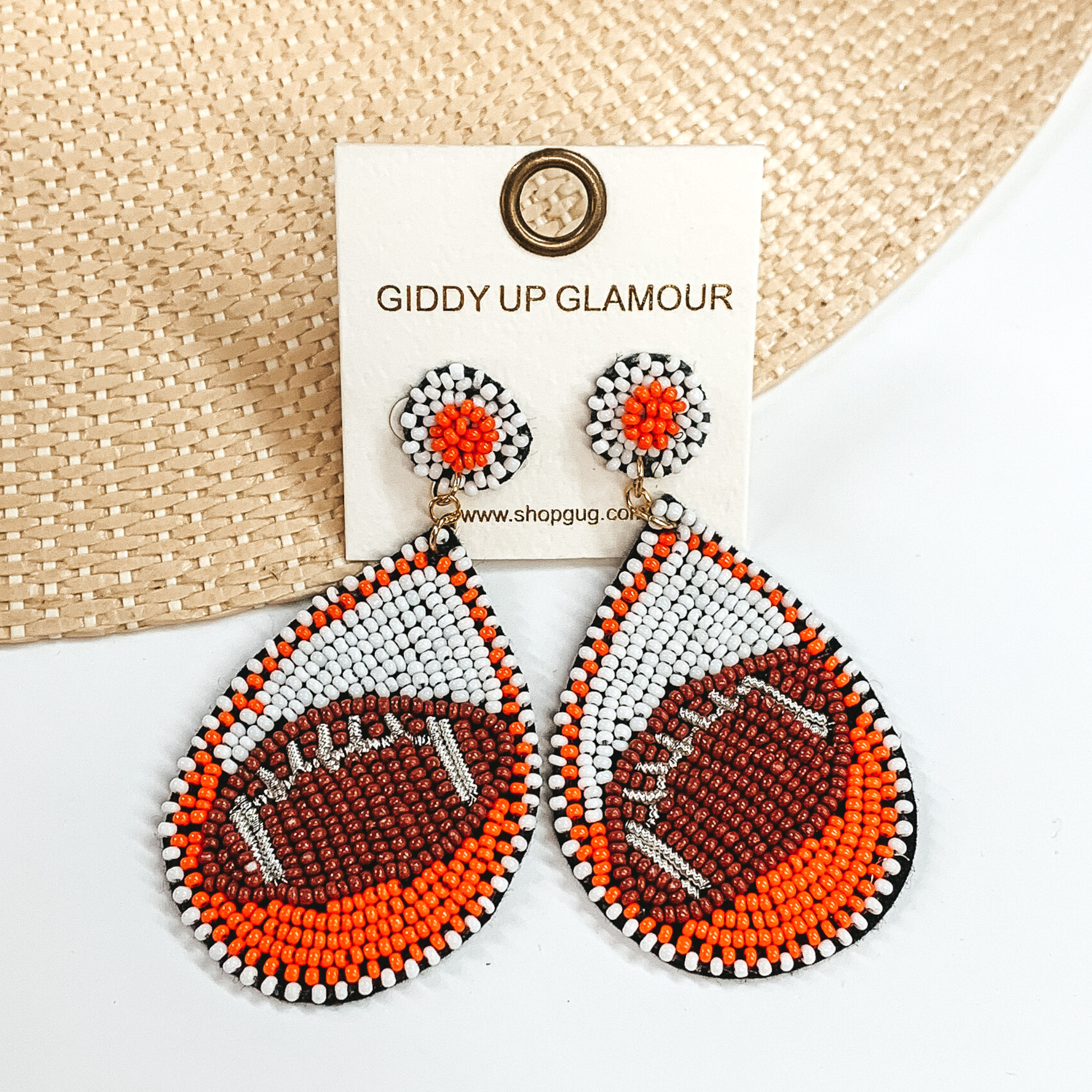 Pictured are a pair of beaded post back earrings with teardrop pendants. These earrings include white and orange beads with a brown football design. These earrings are pictured laying in a straw hat brim on a white background. 