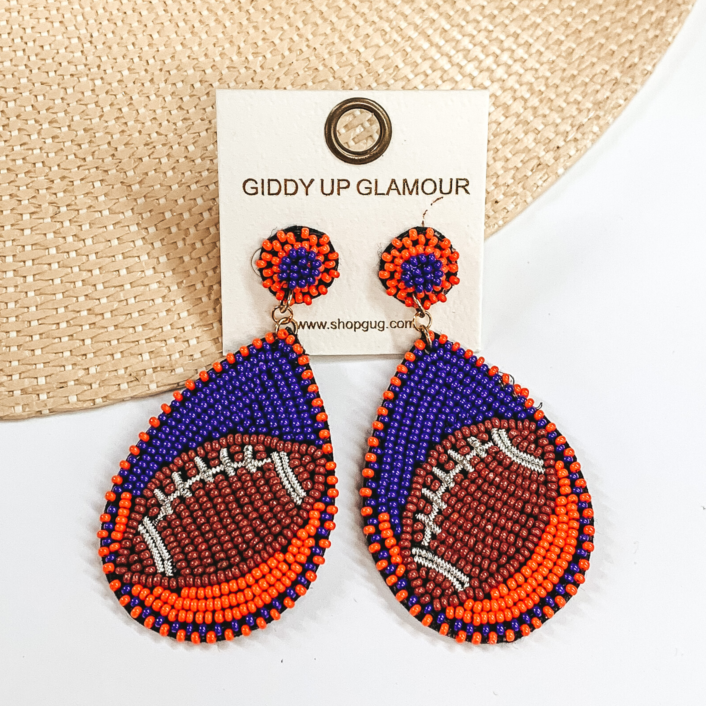 Pictured are a pair of beaded post back earrings with teardrop pendants. These earrings include navy and orange beads with a brown football design. These earrings are pictured laying in a straw hat brim on a white background. 