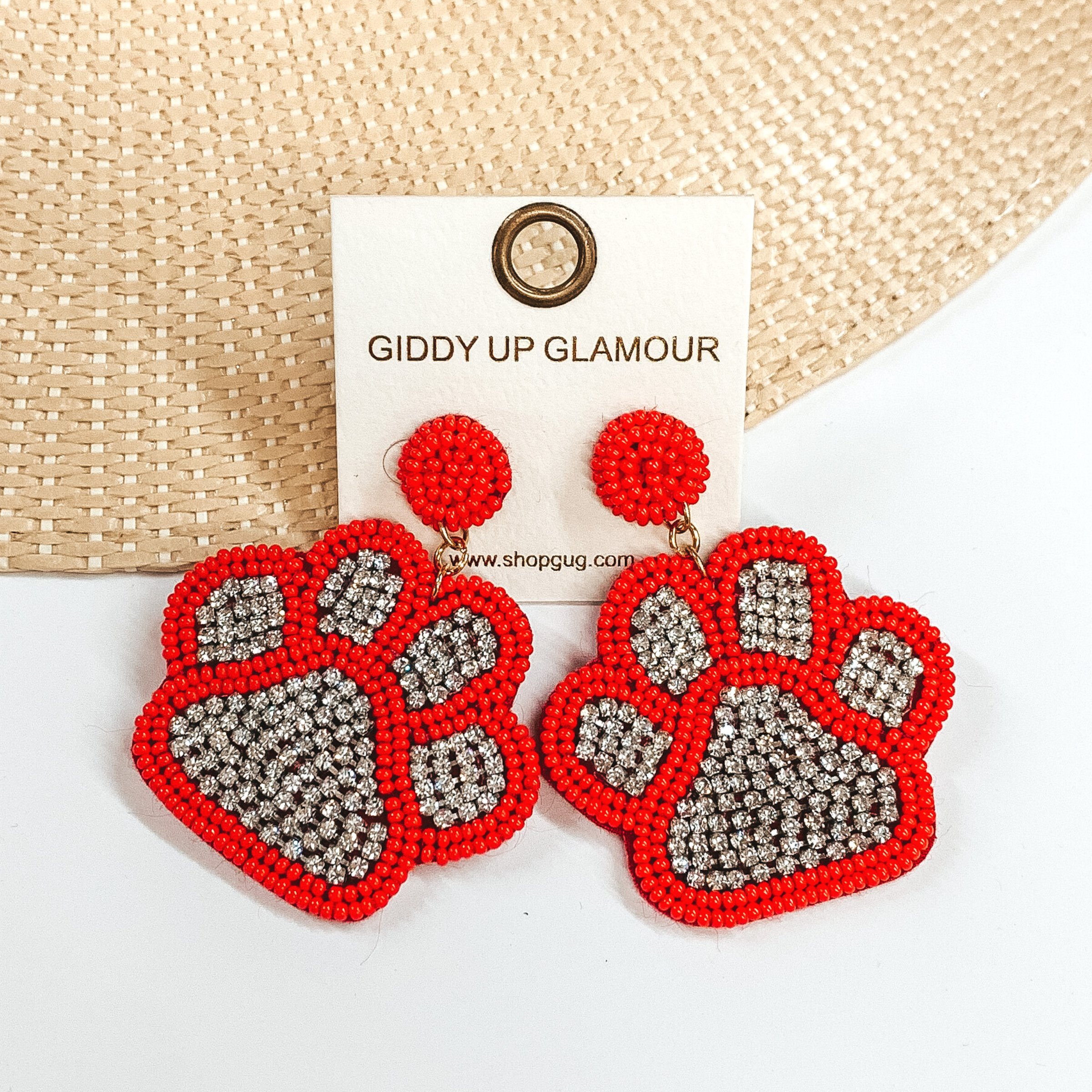 Pictured are a pair of beaded post back earrings with paw print pendants. These earrings include red orange beads and clear crystals in the center. These earrings are pictured laying on a straw hat brim on a white background. 