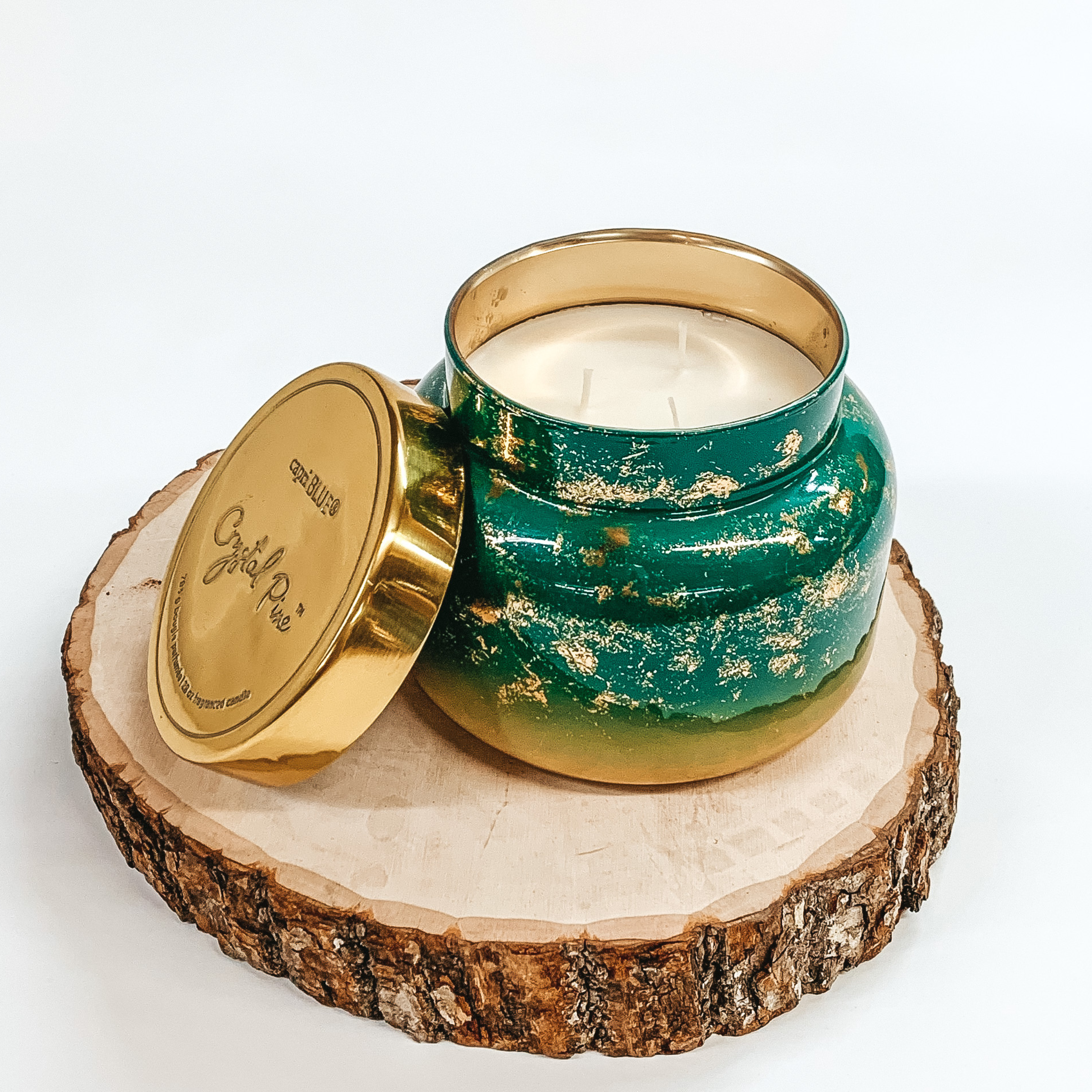 Green ombre candle jar with gold glimmer throughout. This candle is also pictured with a gold lid leaning against the candle. This candle is pictured sitting on a piece of wood on a white background. 