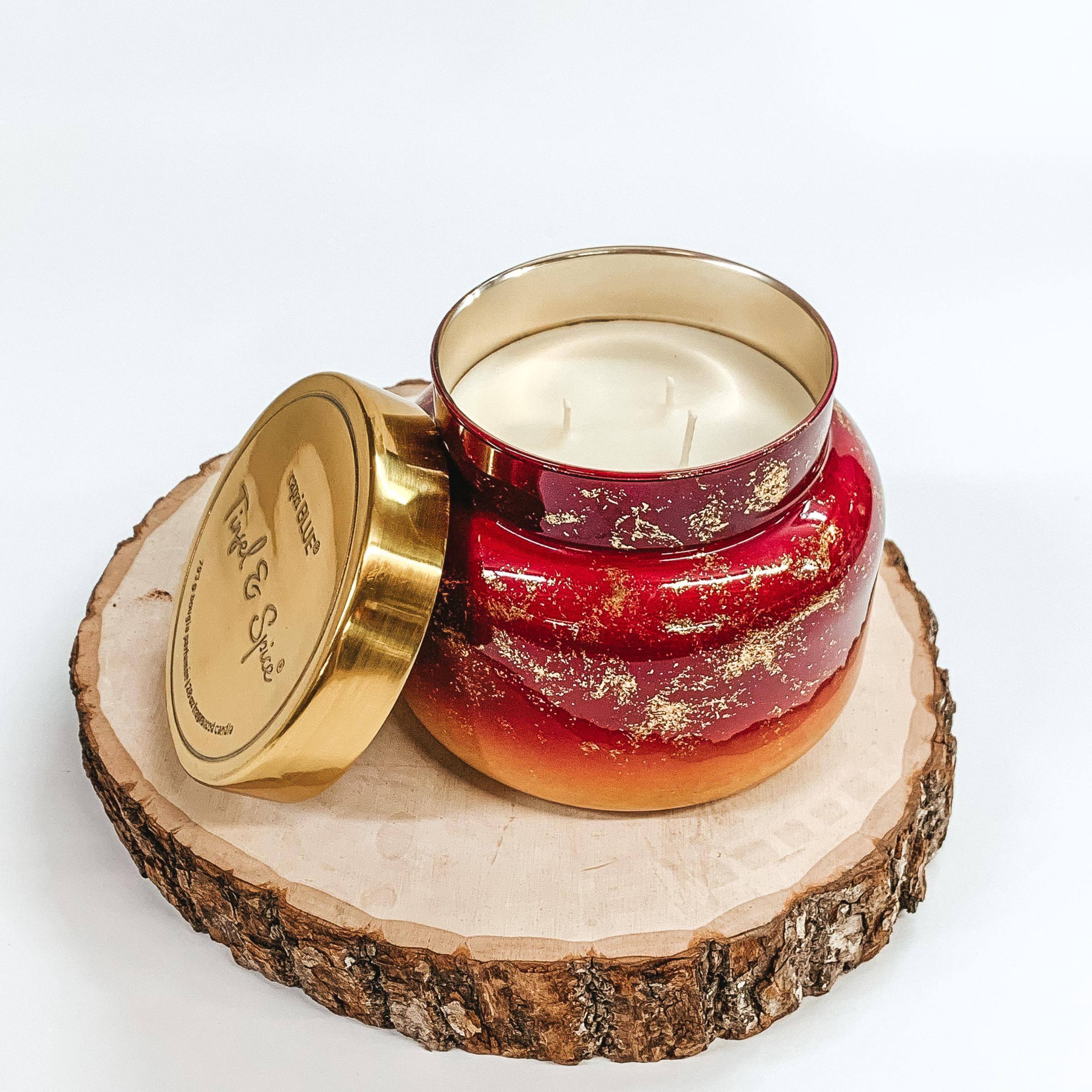 Red ombre candle jar with gold glimmer throughout. This candle is also pictured with a gold lid leaning against the candle. This candle is pictured sitting on a piece of wood on a white background. 