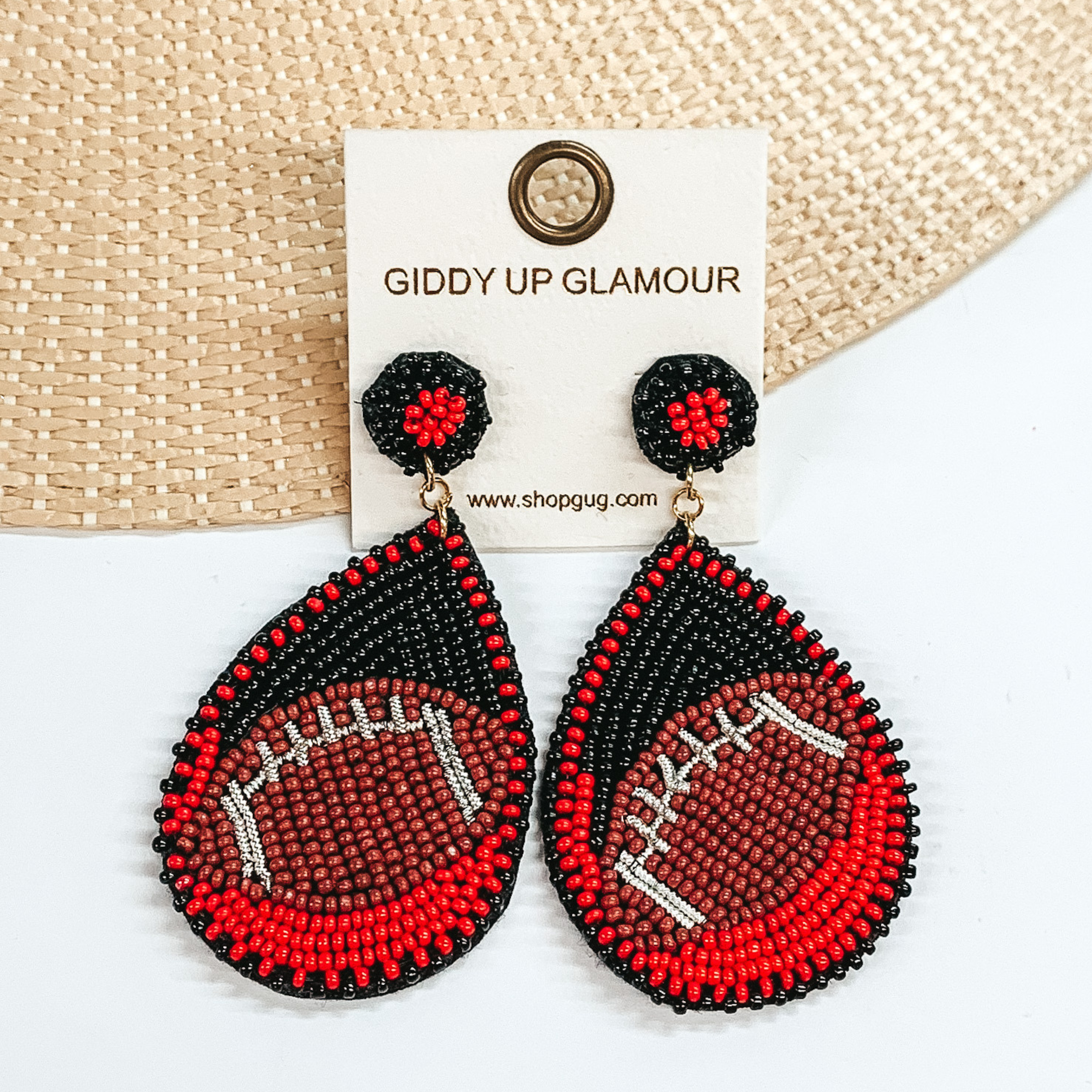 Pictured are a pair of beaded post back earrings with teardrop pendants. These earrings include black and red beads with a brown football design. These earrings are pictured laying in a straw hat brim on a white background.