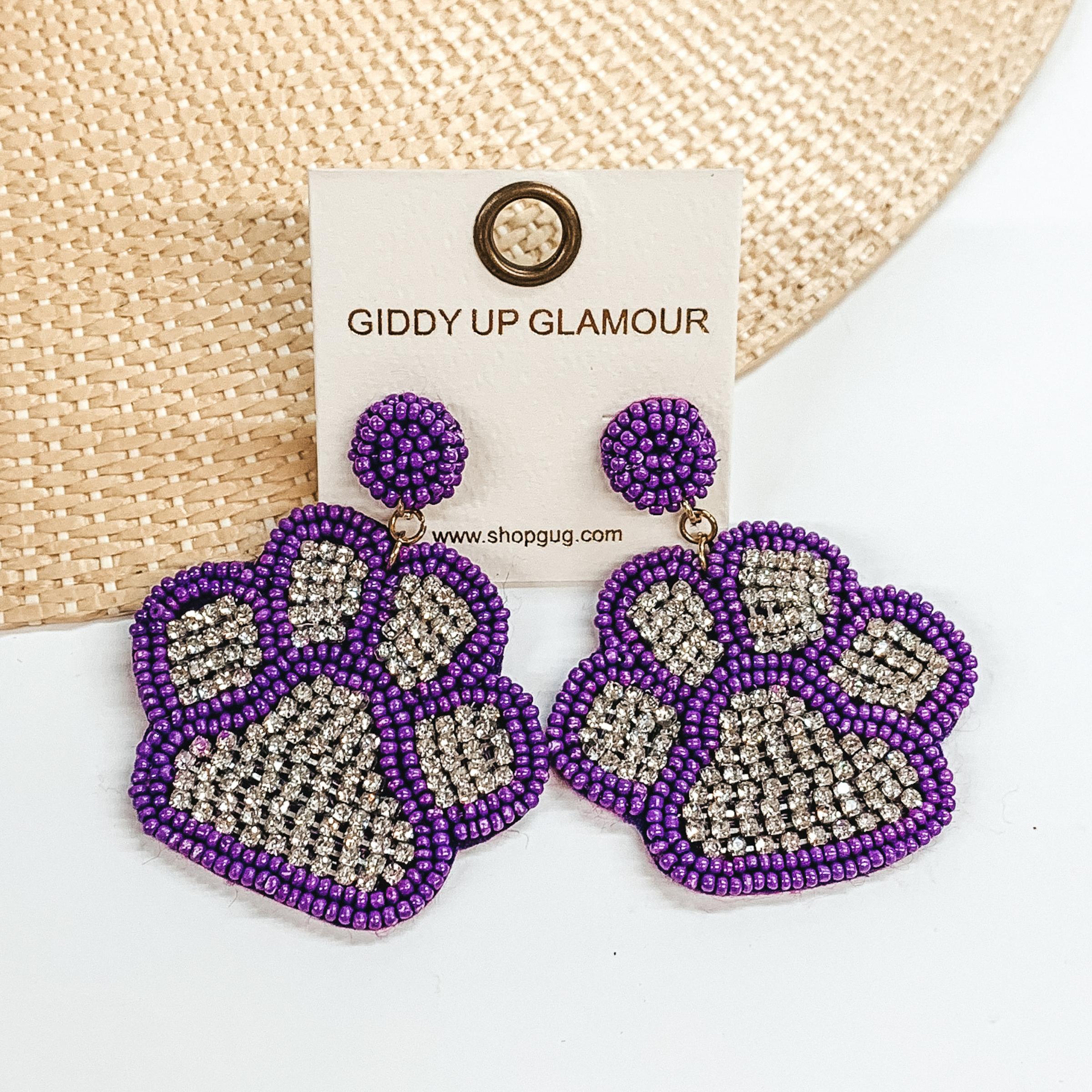 Pictured are a pair of beaded post back earrings with paw print pendants. These earrings include purple beads and clear crystals in the center. These earrings are pictured laying on a straw hat brim on a white background.