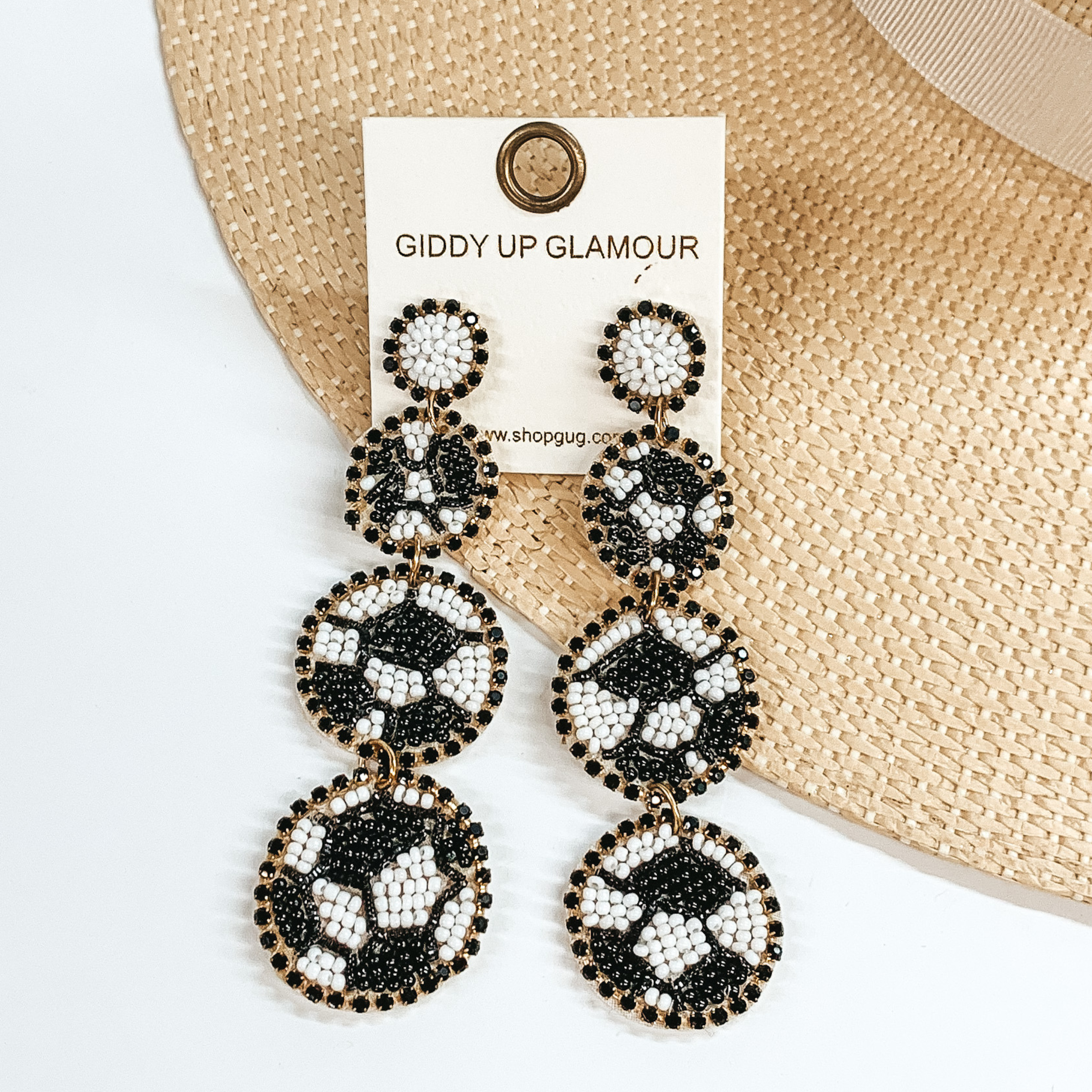 White beaded post back earrings with three beaded soccer ball pendants. These earrings also include a black crystal outline. These earrings are pictured partially laying on a straw hat brim on a white background. 