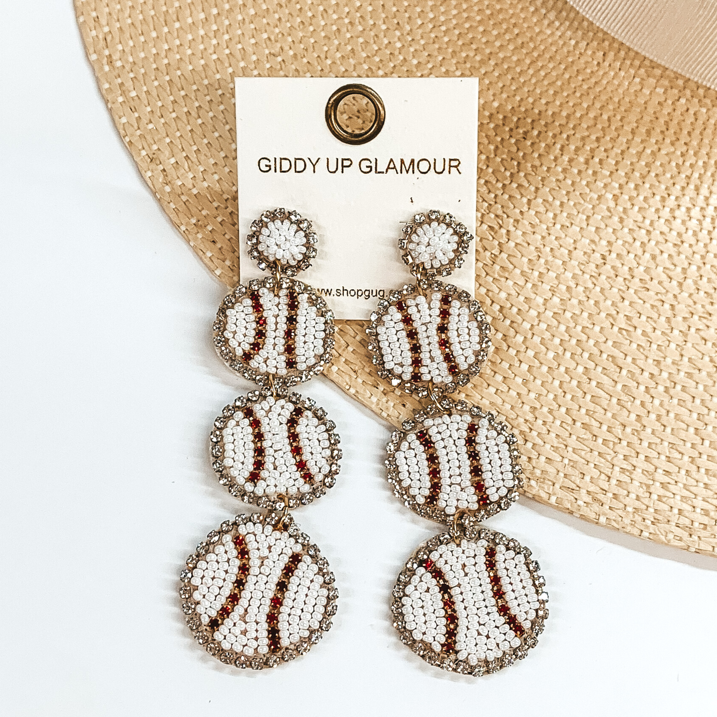 White beaded post back earrings with three beaded baseball pendants. These earrings also include a black crystal outline. These earrings are pictured partially laying on a straw hat brim on a white background. 