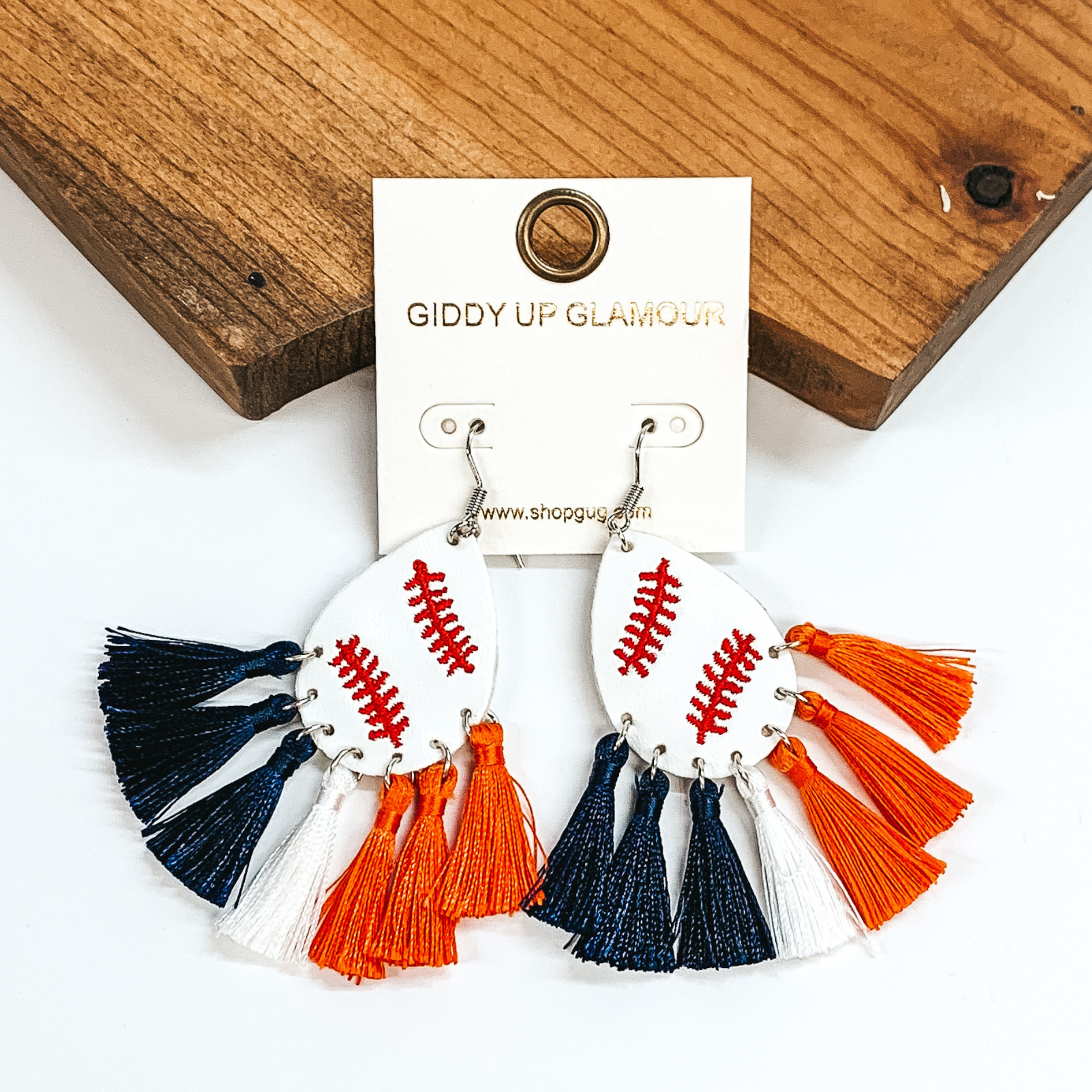 Pictured are a pair of silver fish hook earrings with a white teardrop with red stitching to look like a baseball. These earrings also include navy, white, and orange tassels at the bottom. These earrings are pictured in front of a brown block on a white background. 