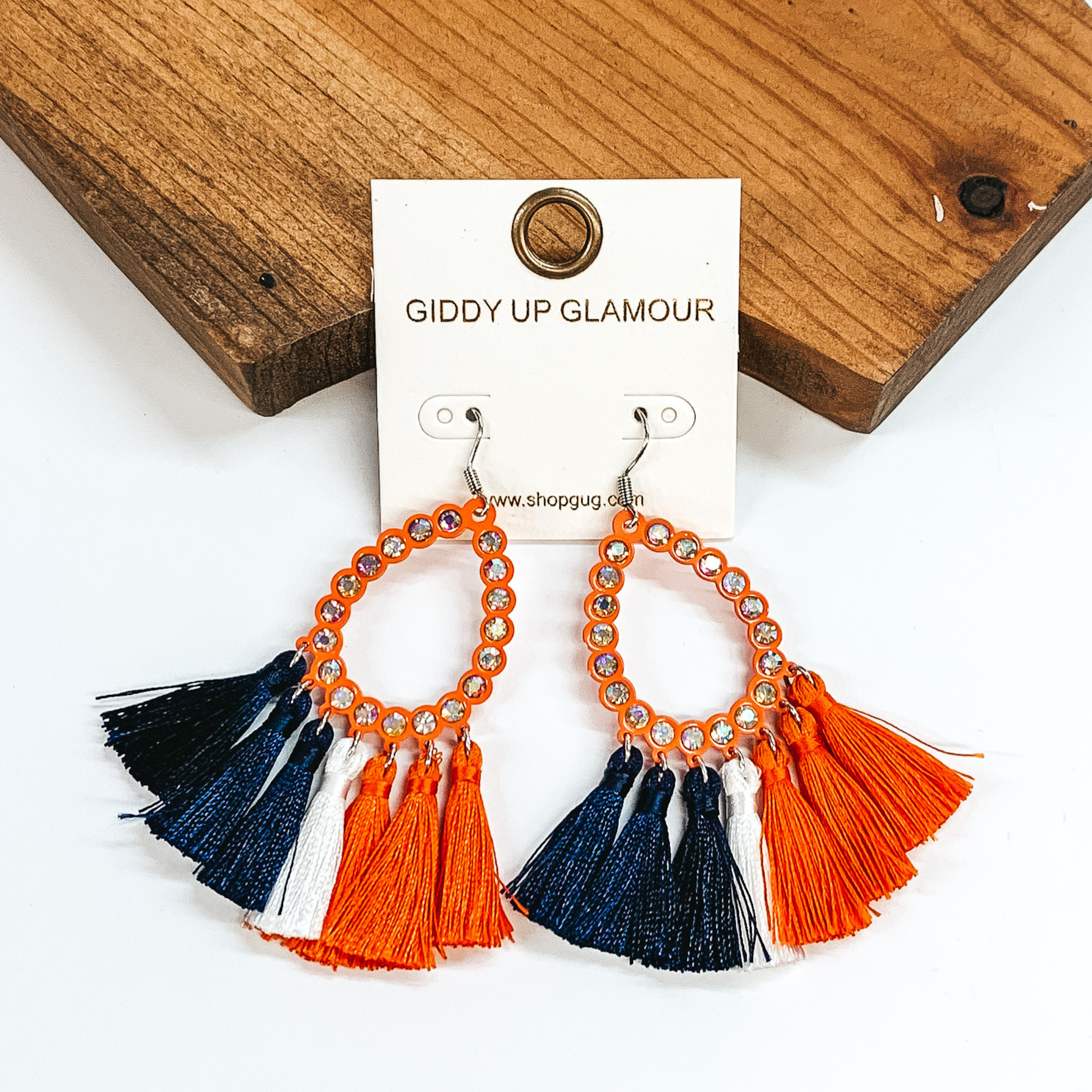 Pictured are a pair of silver fish hook earrings with an open, orange teardrop pendant with ab crystals. These earrings also include navy, white, and orange tassels at the bottom. These earrings are pictured in front of a brown block on a white background. 