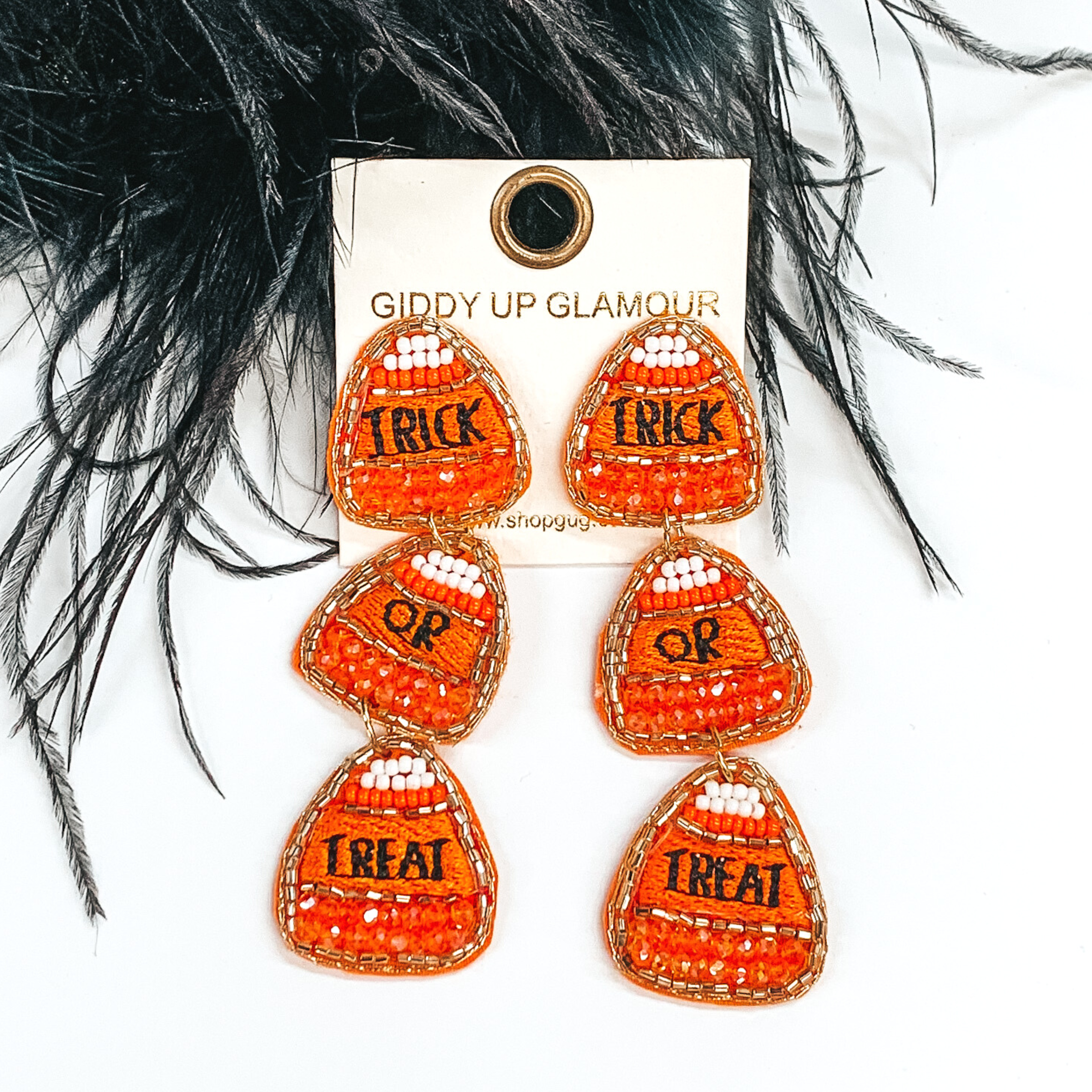 Three tiered beaded candy corn earrings. These earrings include one word stitched in black on each candy corn, the words are "trick," "or," and "treat." These earrings are pictured on a white background with black feathers at the top. 
