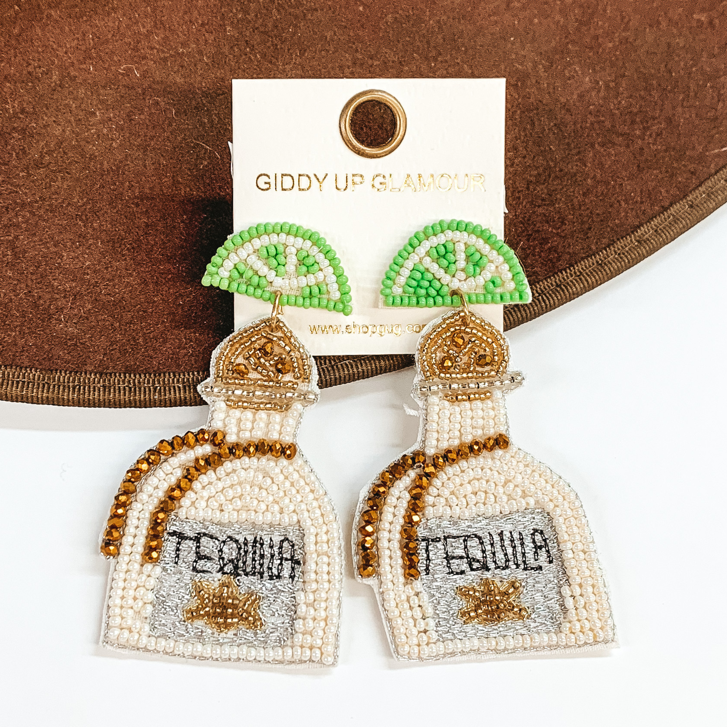Light green beaded lime stud earrings with hanging beaded bottle. The bottle has a gold beaded top with a ivory beaded body with brown beaded stripes. The center on the bottom is grey stitched with the word "TEQUILA" stitched in black. These earrings are pictured on a dark brown object on a white background.