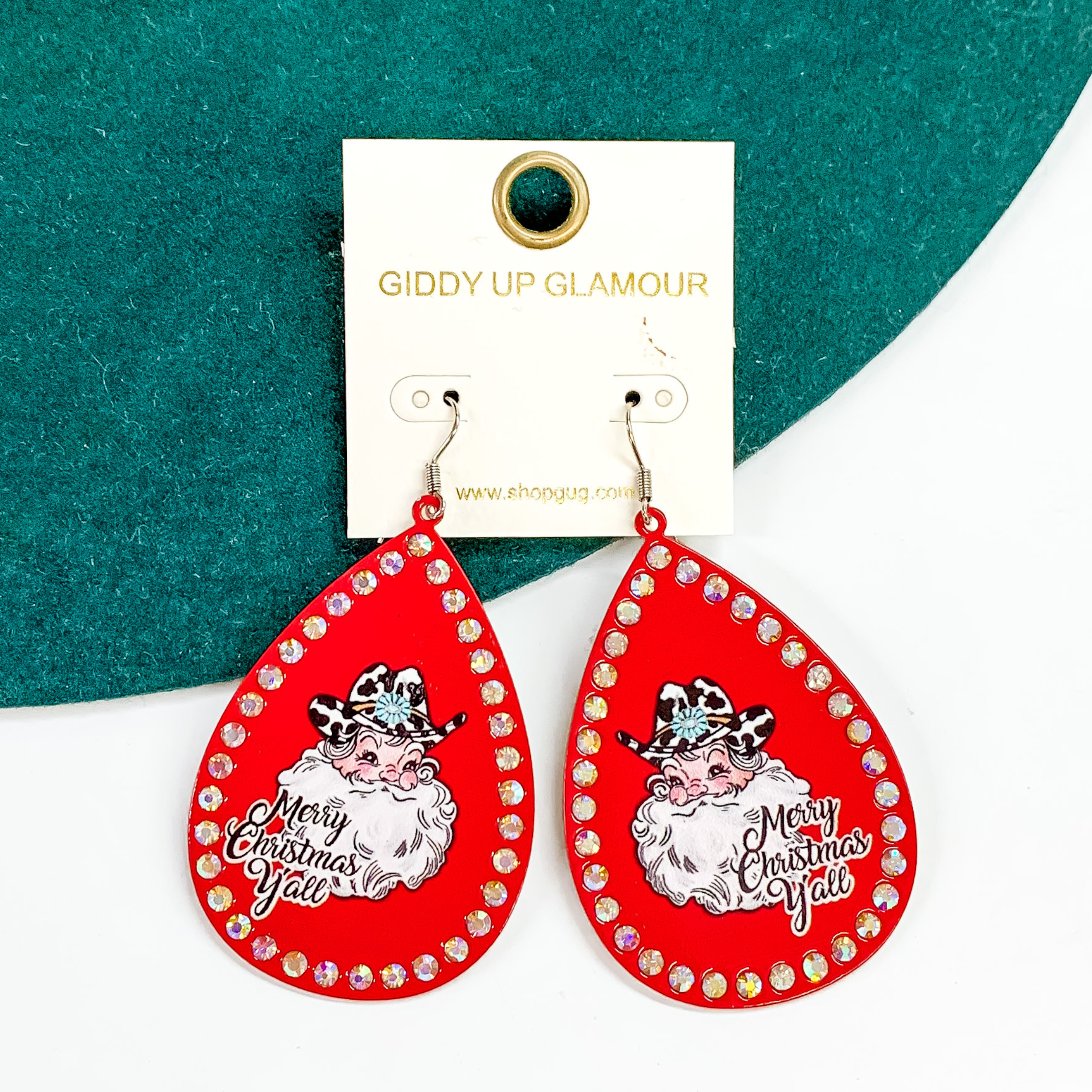 Teardrop dangle earrings with a red design with an AB crystal outline. These earrings also include a printed Santa in the middle with a white and black cow print hat that has a concho belt. The words "Merry Christmas Y'all" is under the Santa head. These earrings are pictured partially laying on a green felt hat on a white background.