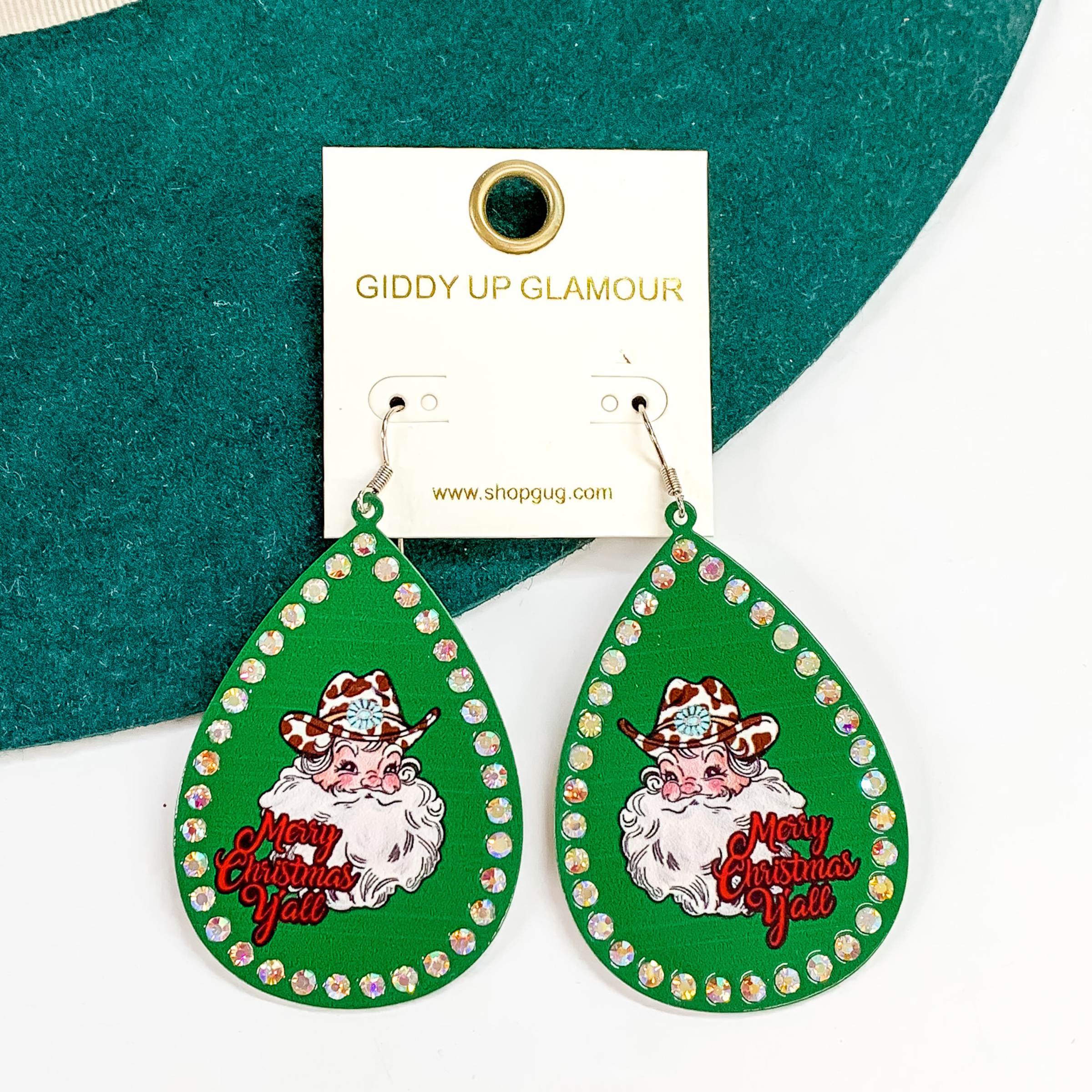 Teardrop dangle earrings with a green design with an AB crystal outline. These earrings also include a printed Santa in the middle with a white and brown cow print hat that has a concho belt. The words "Merry Christmas Y'all" is under the Santa head. These earrings are pictured partially laying on a green felt hat on a white background.